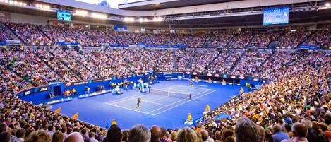 A large indoor tennis stadium with multiple tiers of seating, a central court, and a scoreboard.