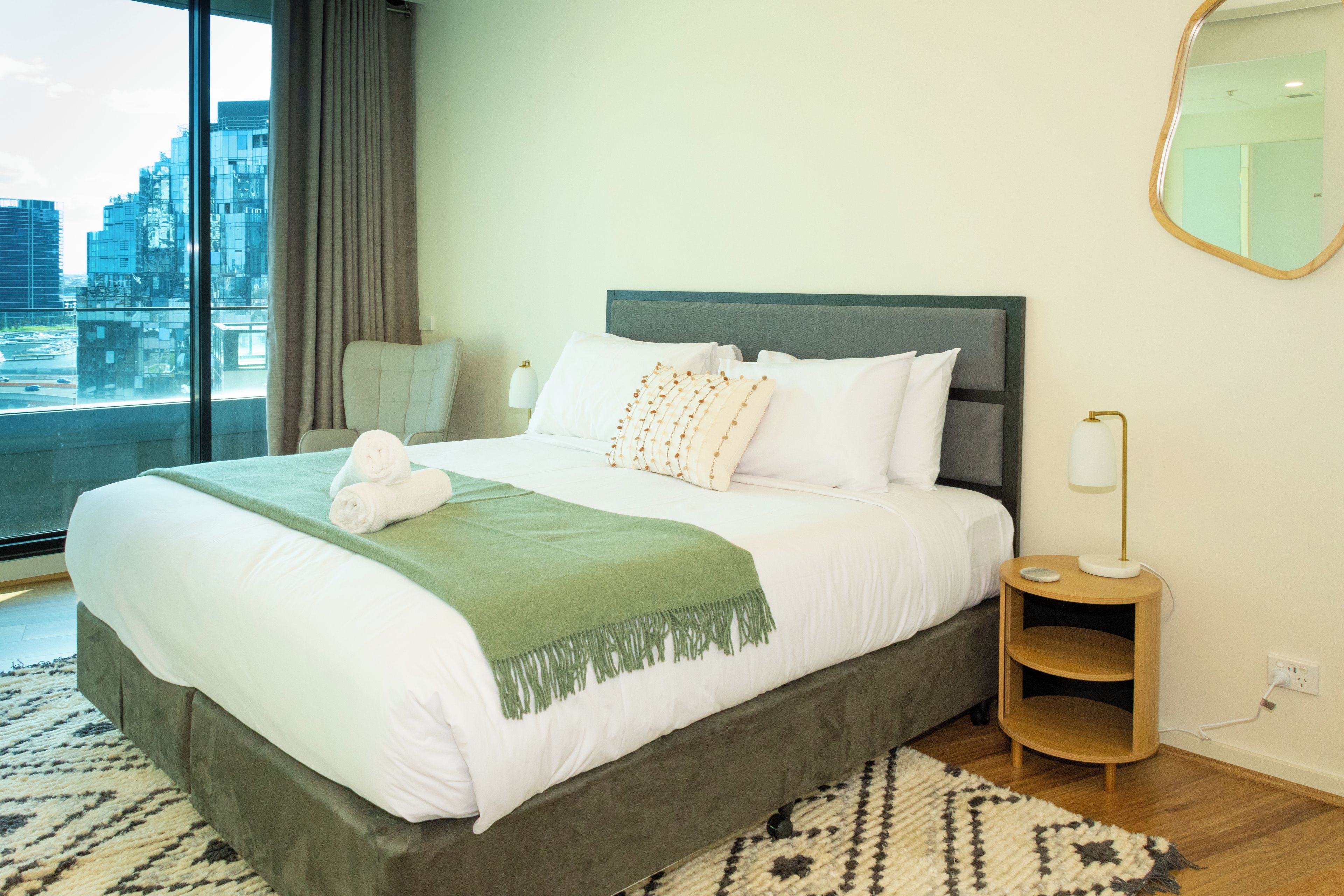 A hotel room with a large bed, a view of the city through a window, and a bedside table with a lamp.