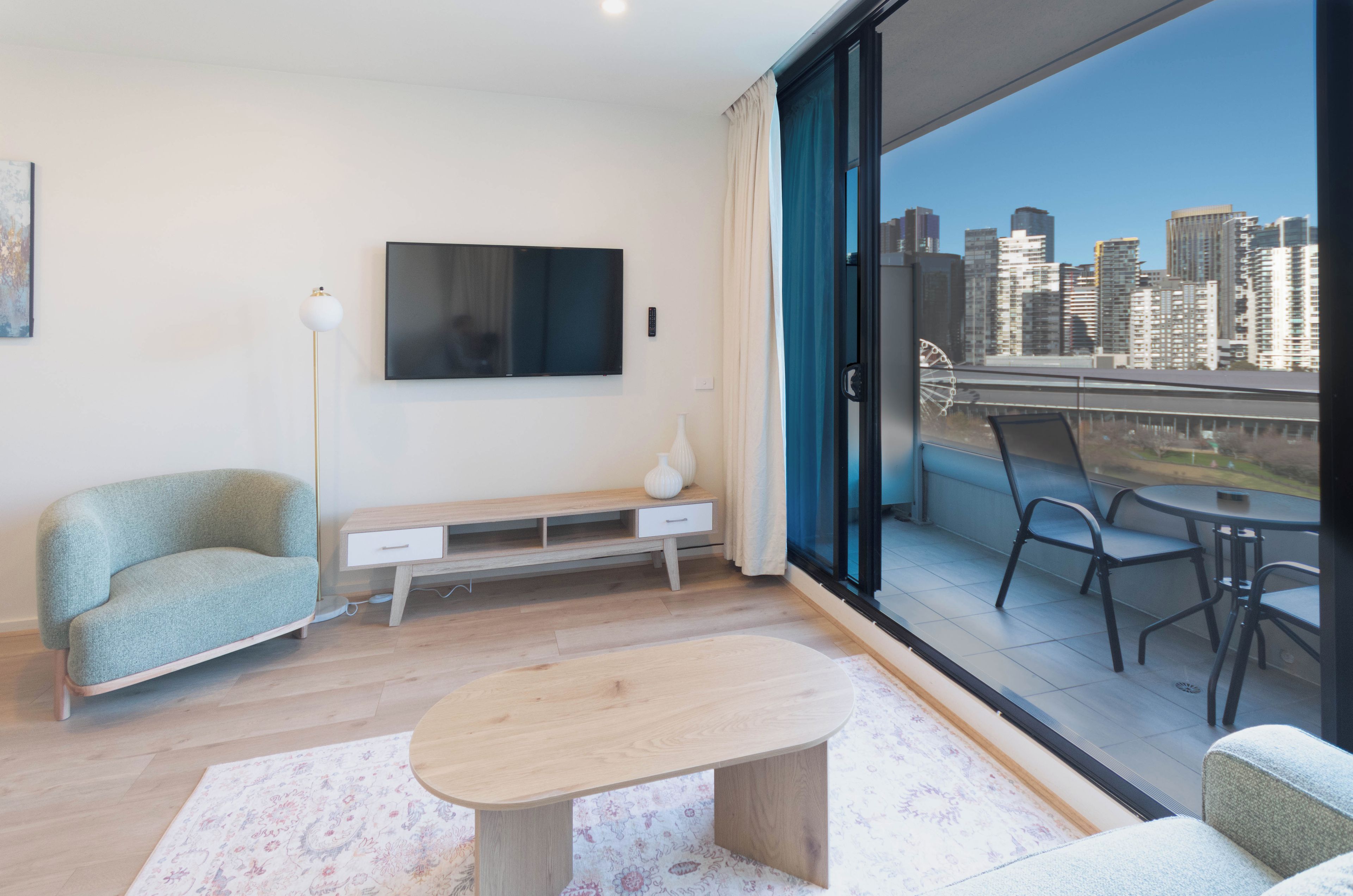 A modern living room with a flat-screen TV, a sofa, a coffee table, and a balcony with a city view.
