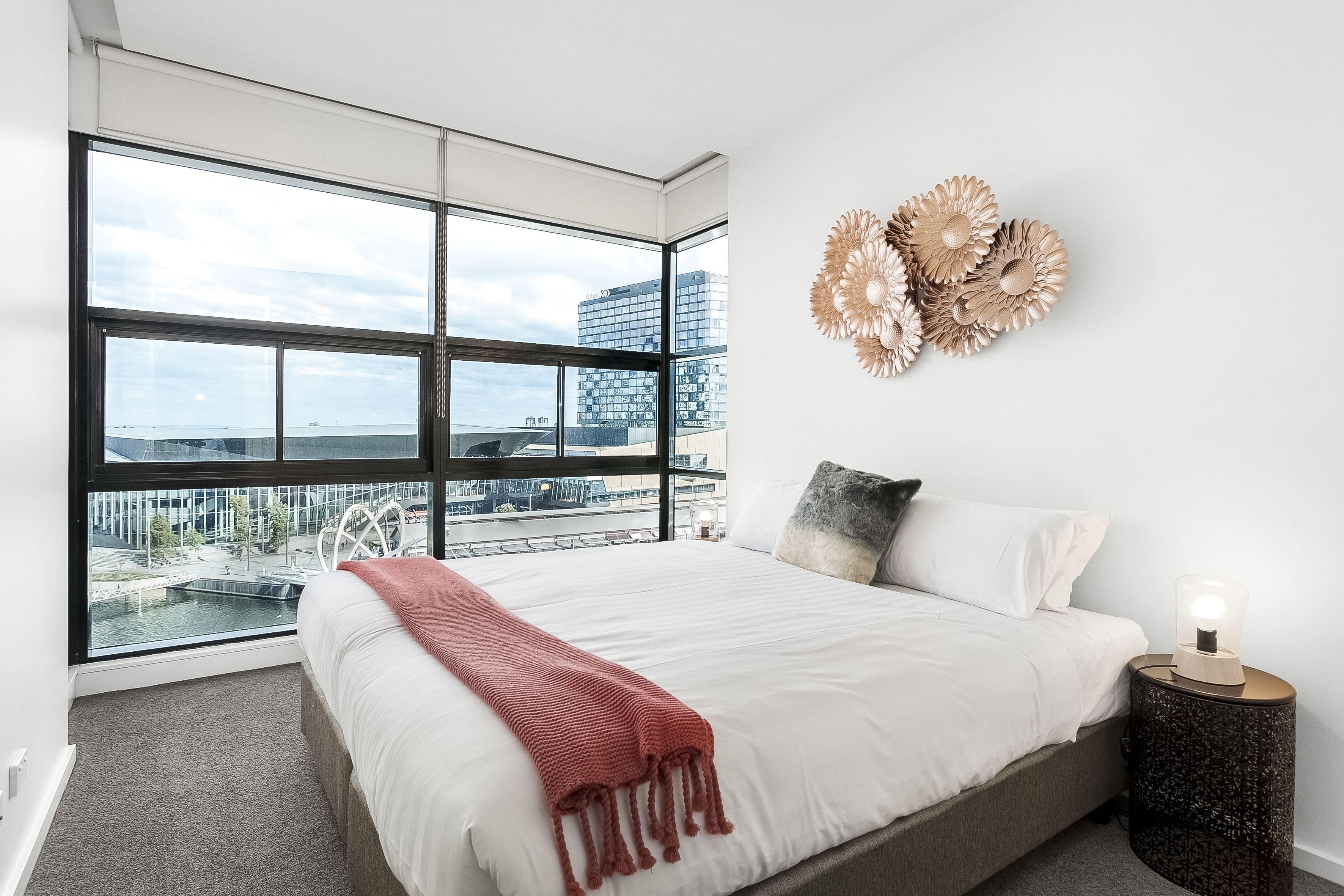 A modern bedroom with a large window, a bed with white bedding, a red throw blanket, decorative wall art, and a bedside lamp.