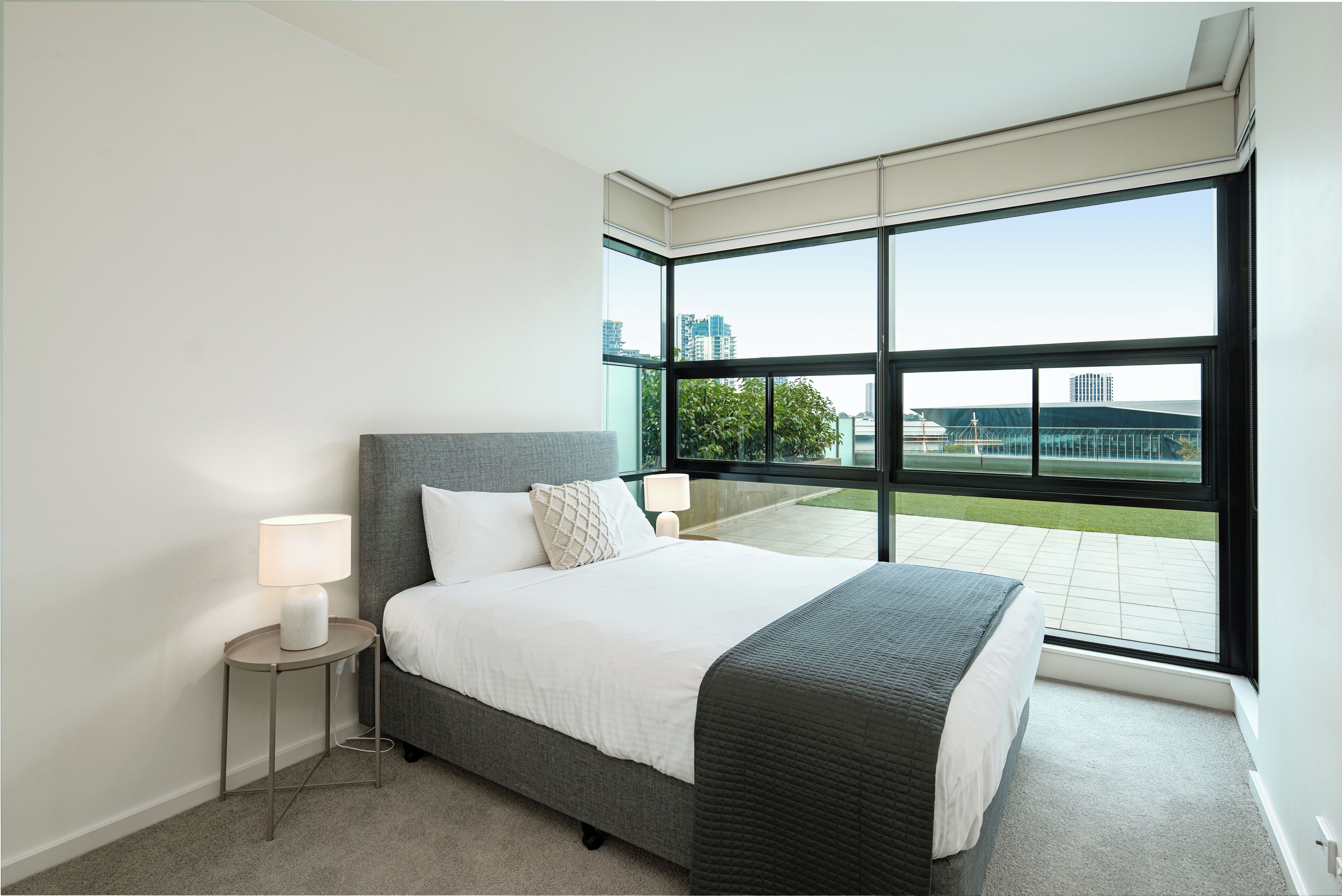 A modern hotel room with a large window, a bed, a bedside table with a lamp, and a view of the city.