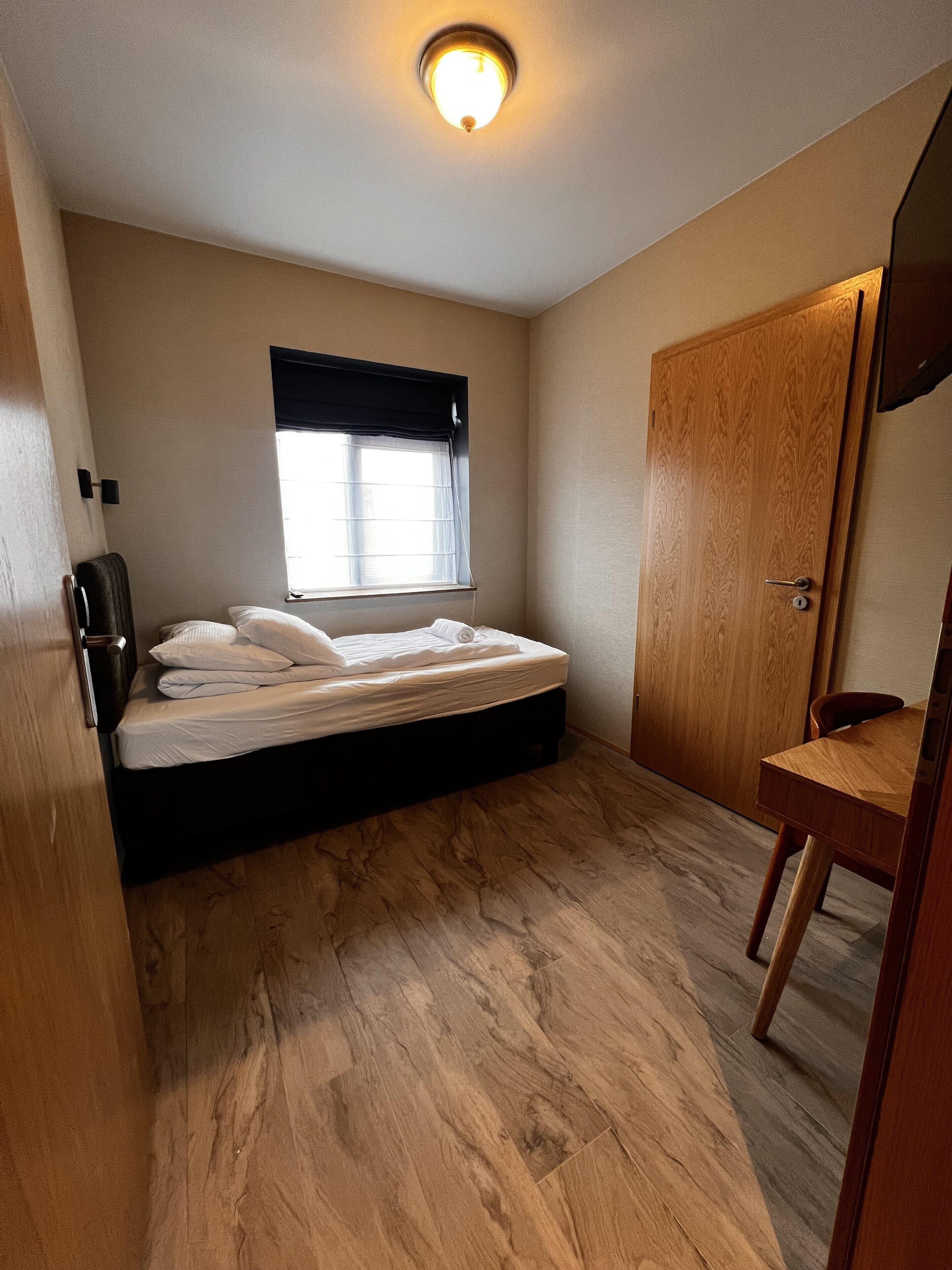 Una habitación pequeña con cama, ventana con persianas, puerta de madera y una mesita.