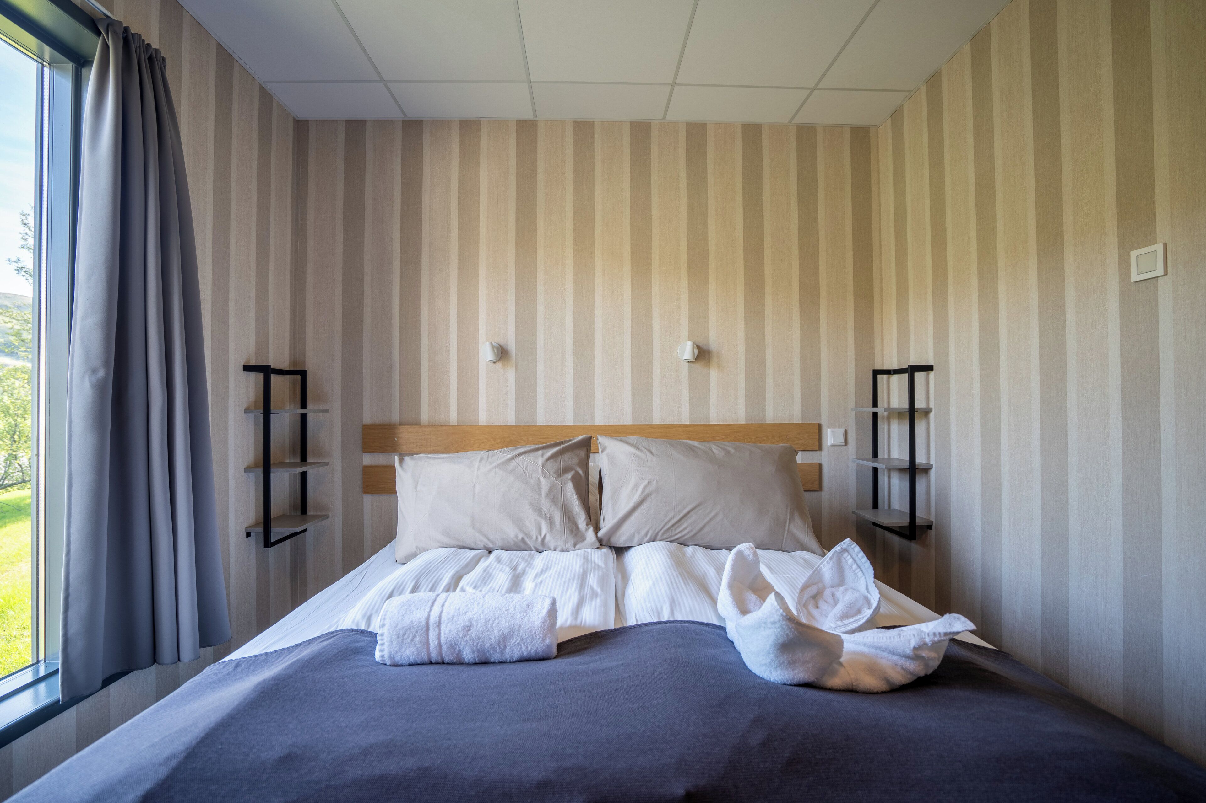 Una cama bien hecha con almohadas y una toalla enrollada, un cabecero de madera y una ventana con cortinas.