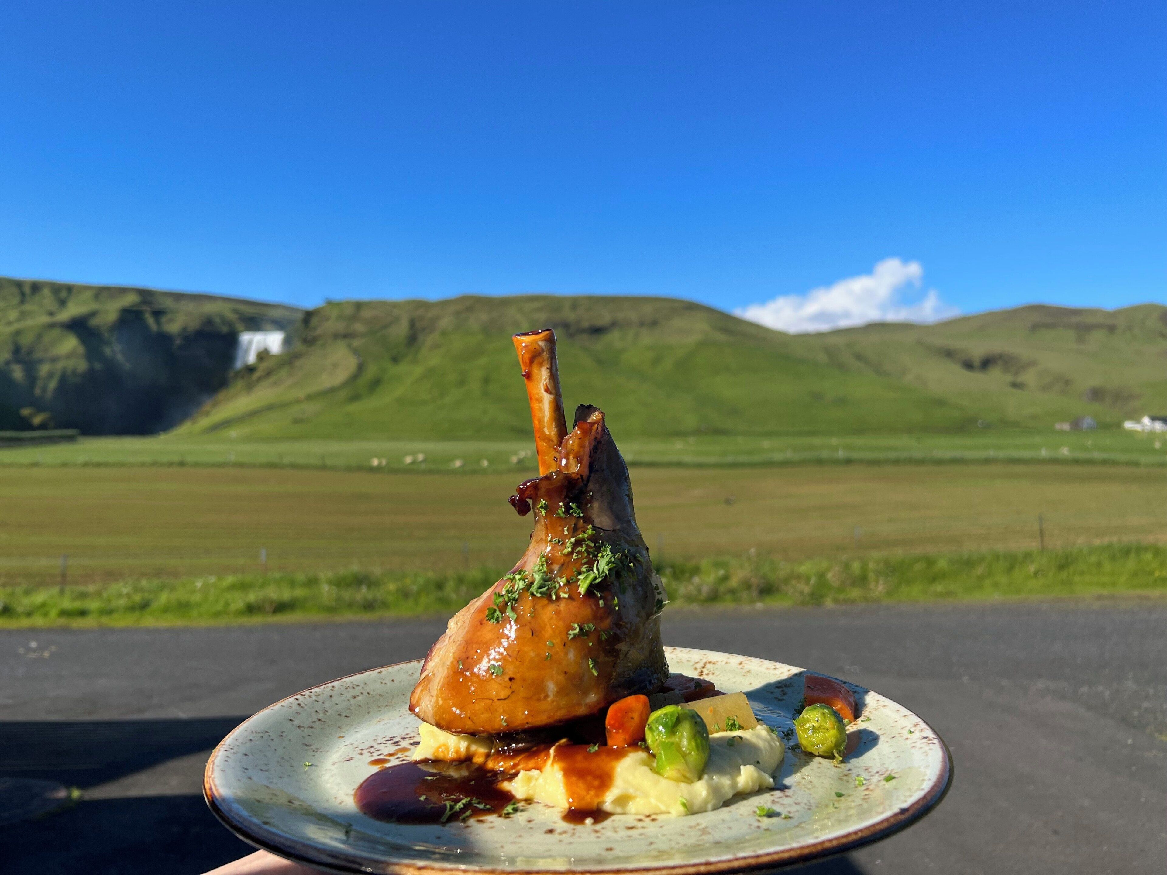 Un plato de cordero con verduras, con un paisaje natural de fondo.