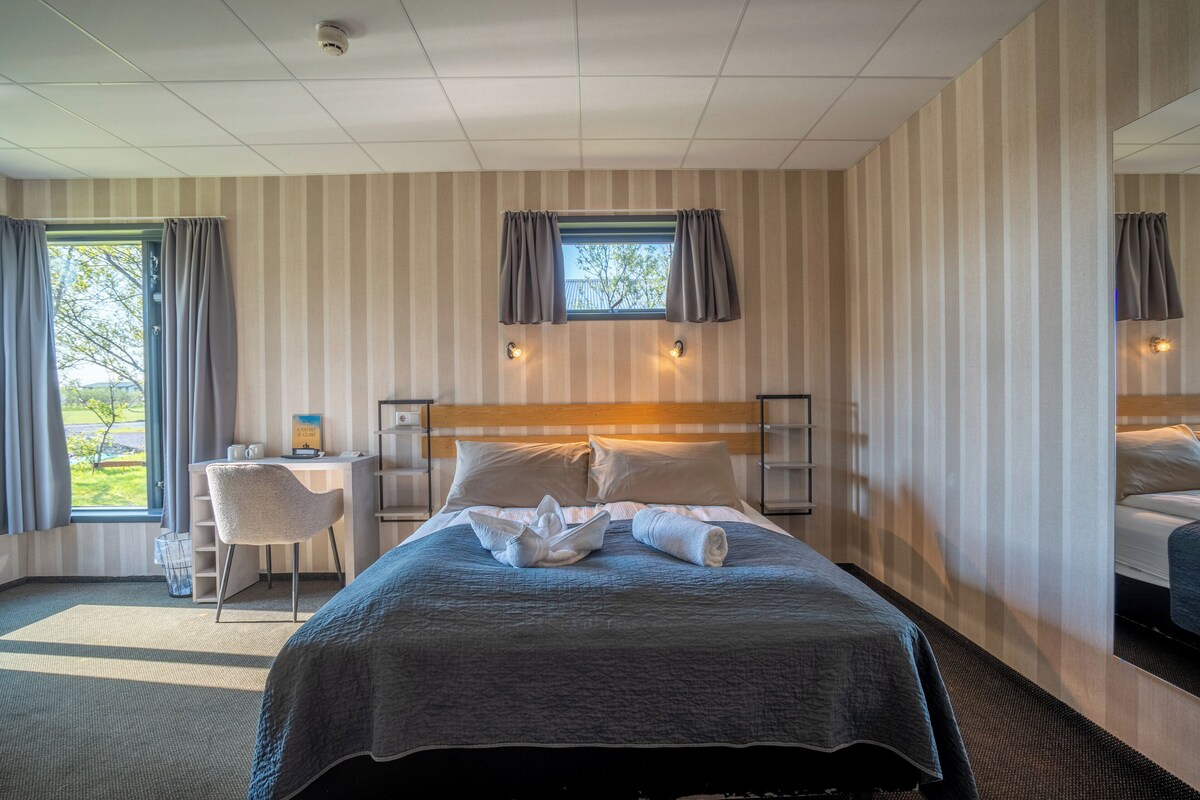 Habitación de hotel con una cama grande, un escritorio con silla, una ventana con cortinas y vistas al exterior.