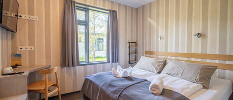 A hotel room with a bed, a desk, a chair, a TV mounted on the wall, and a window with curtains.