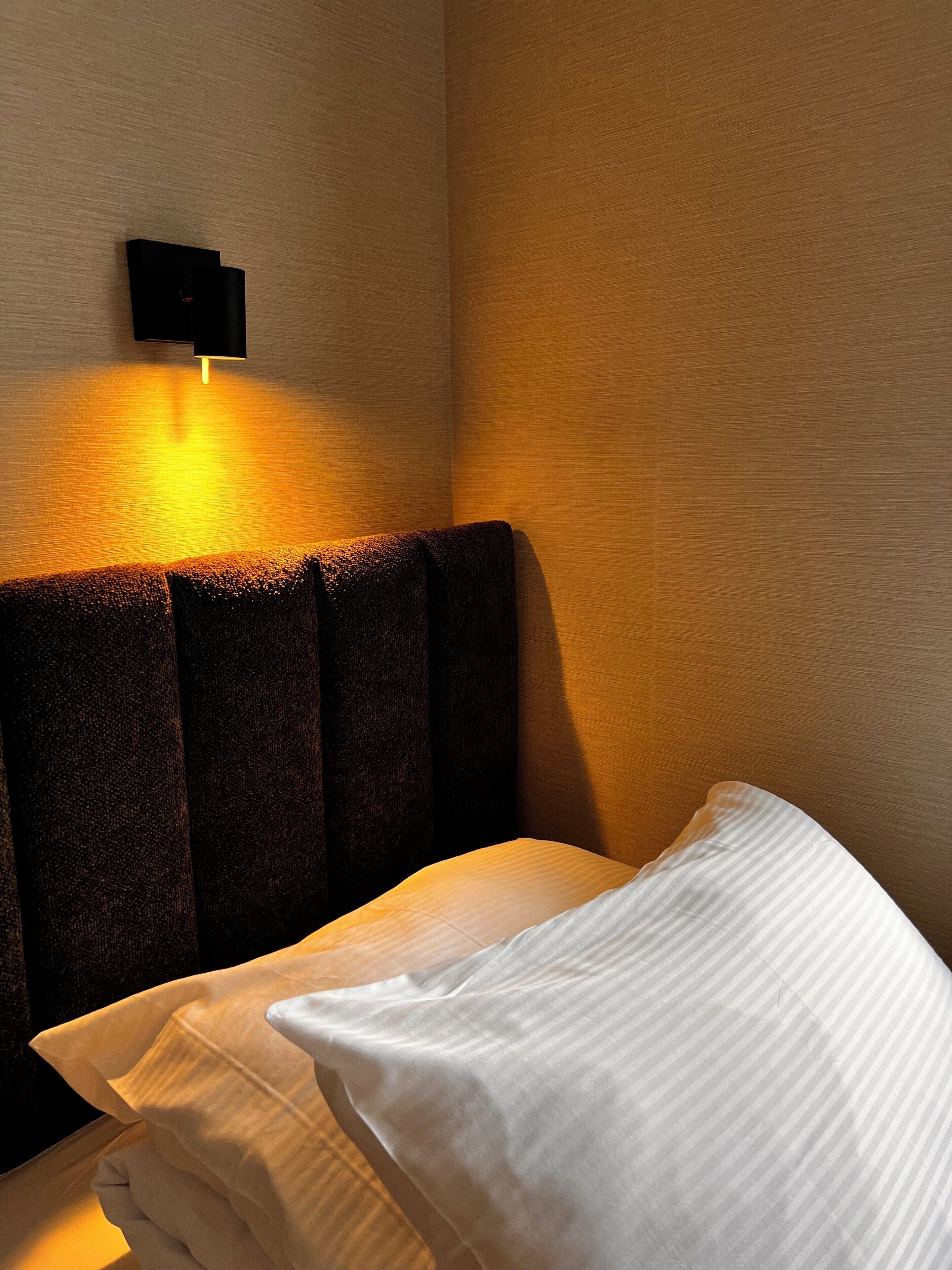 A hotel room with a headboard, a wall-mounted light, and a neatly made bed with pillows.