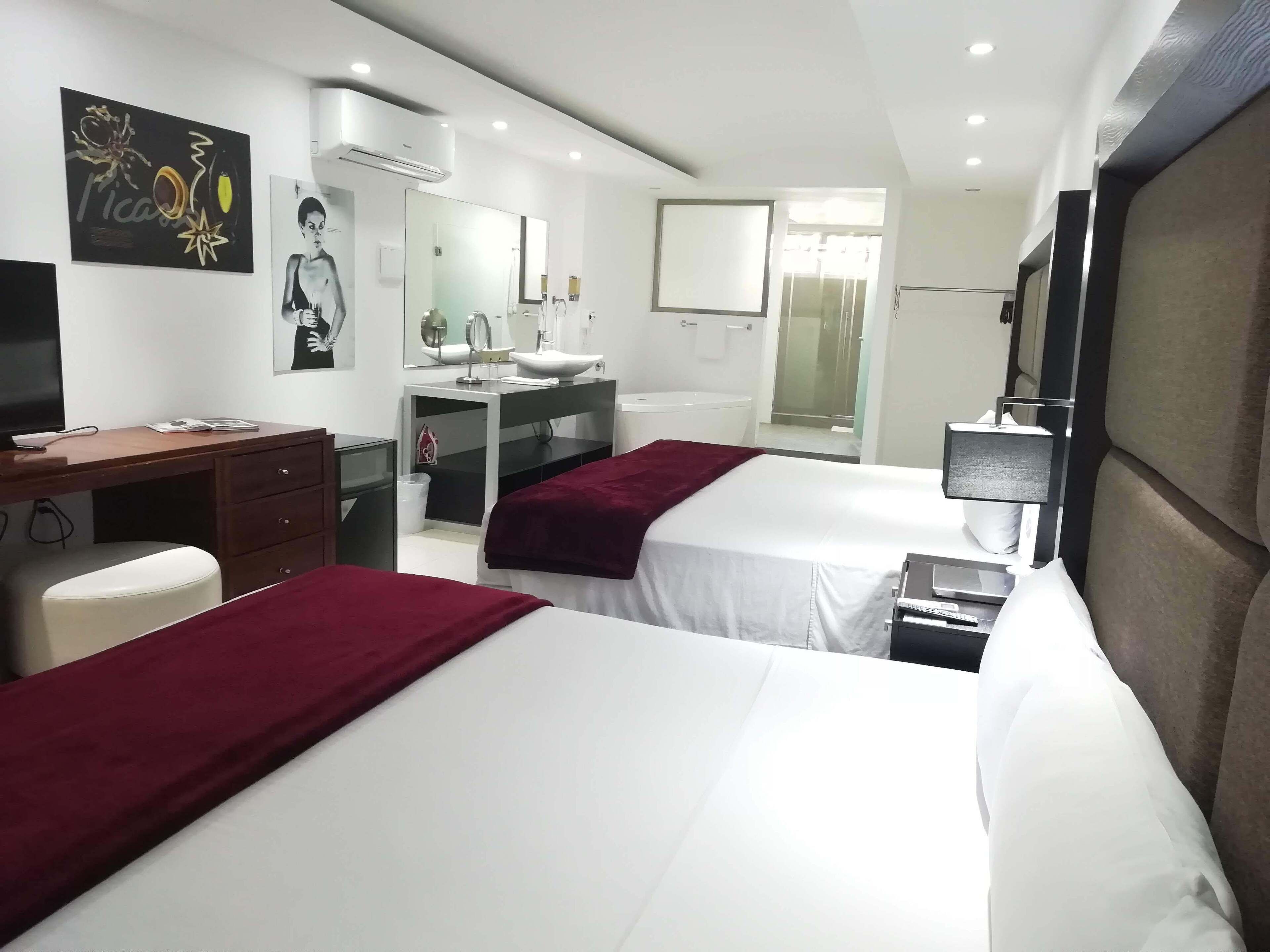 A hotel room with two beds, a desk, a mirror, a sink, and a shower area.