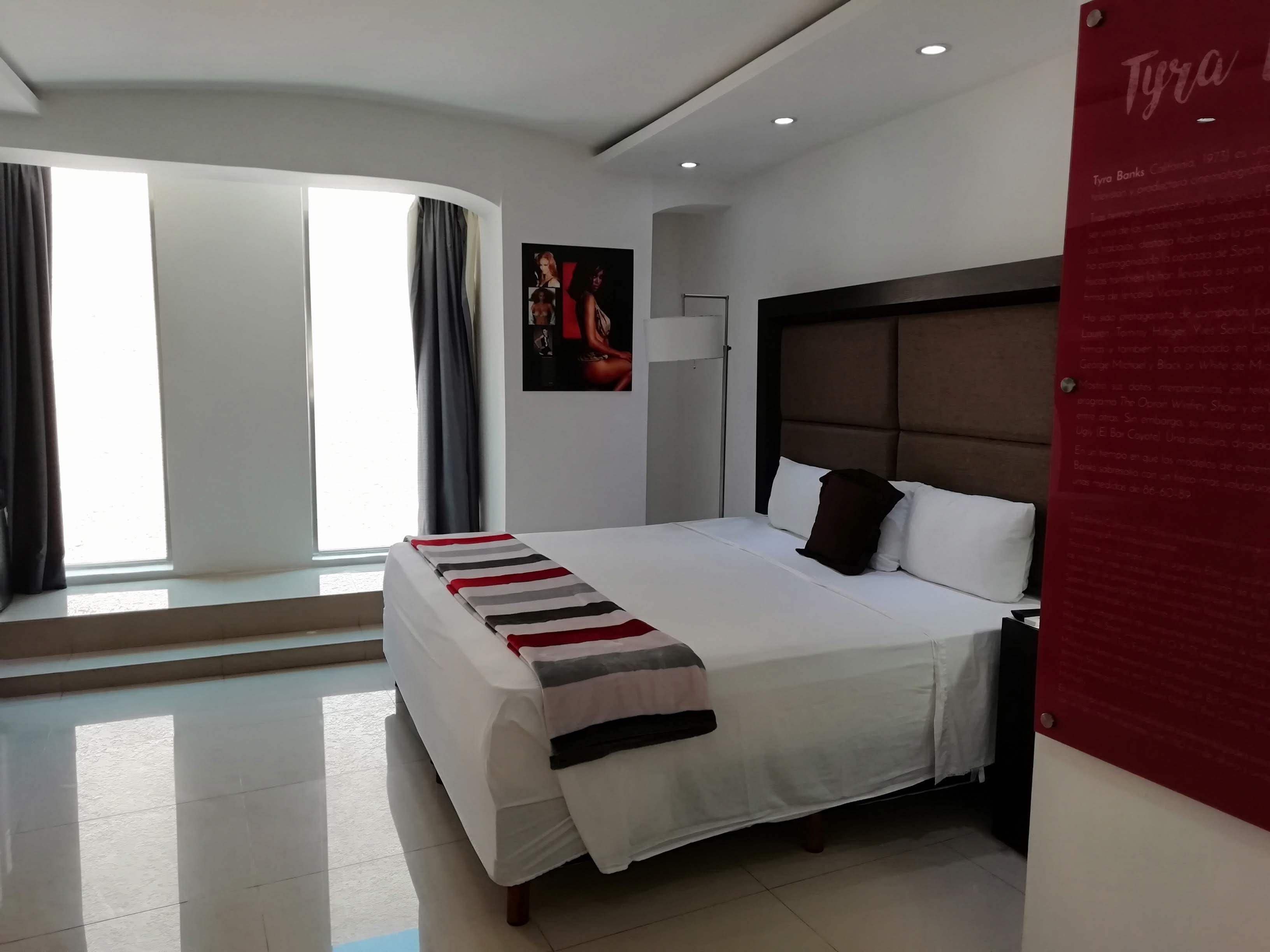 A modern hotel room with a large bed, a headboard, a nightstand, and a window with curtains.