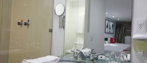 A modern bathroom with a glass shower enclosure, a sink with a mirror, and a bed visible in the background.