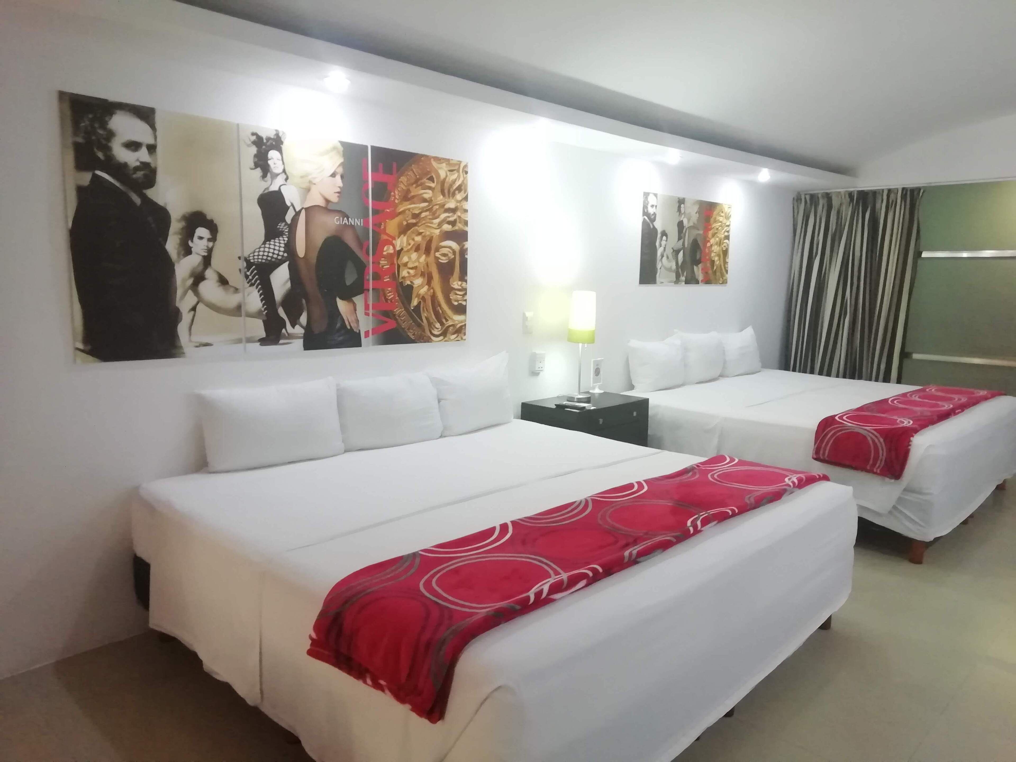 A hotel room with two beds, white bedding, red throw blankets, a nightstand with a lamp, and artwork on the walls.