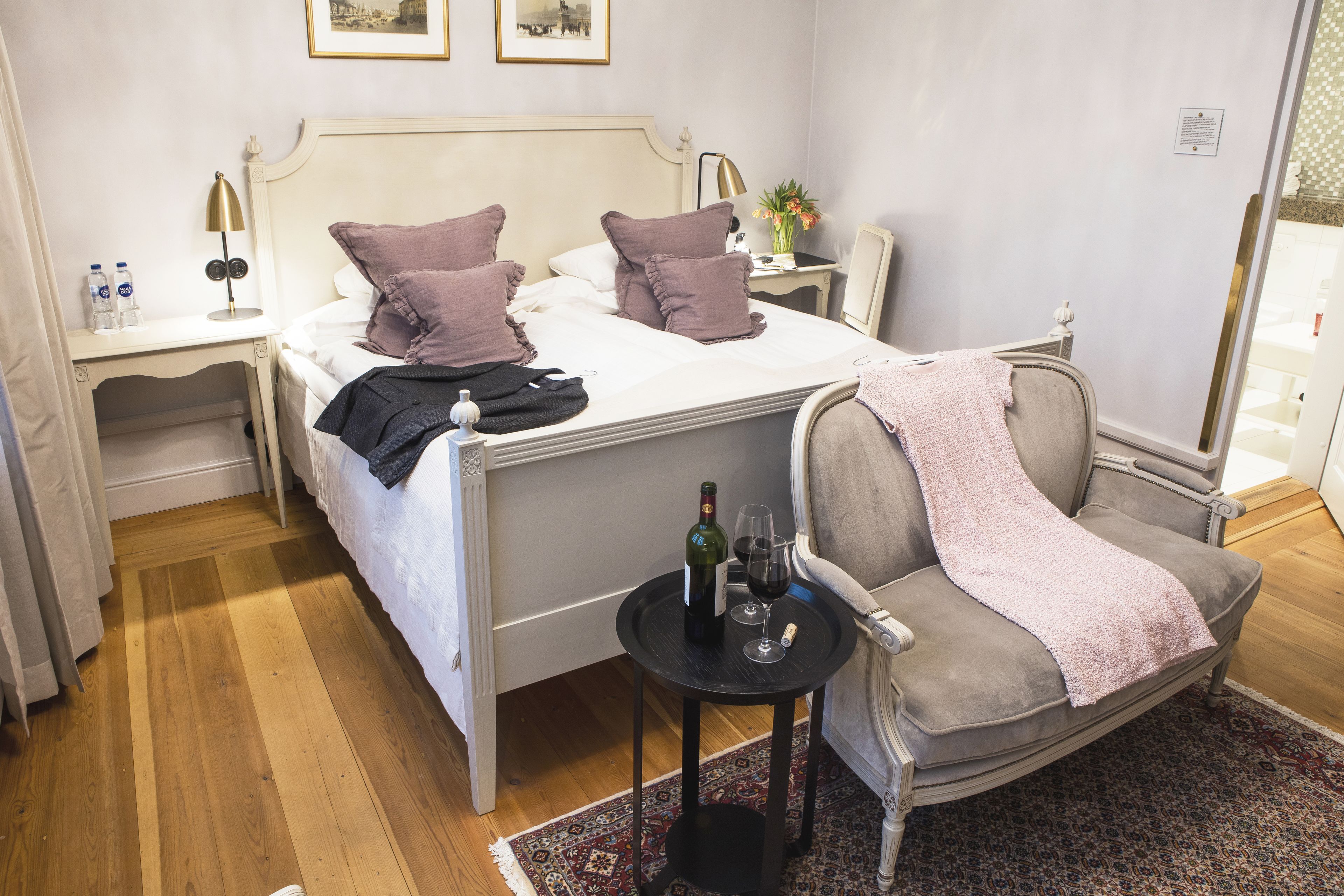 A neatly made bed with pillows, a bedside table with a lamp, a chair with a blanket, and a small table with a bottle and glass.