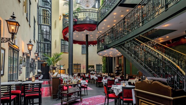 Un restaurante con varias mesas, sillas tapizadas en rojo y una escalera.