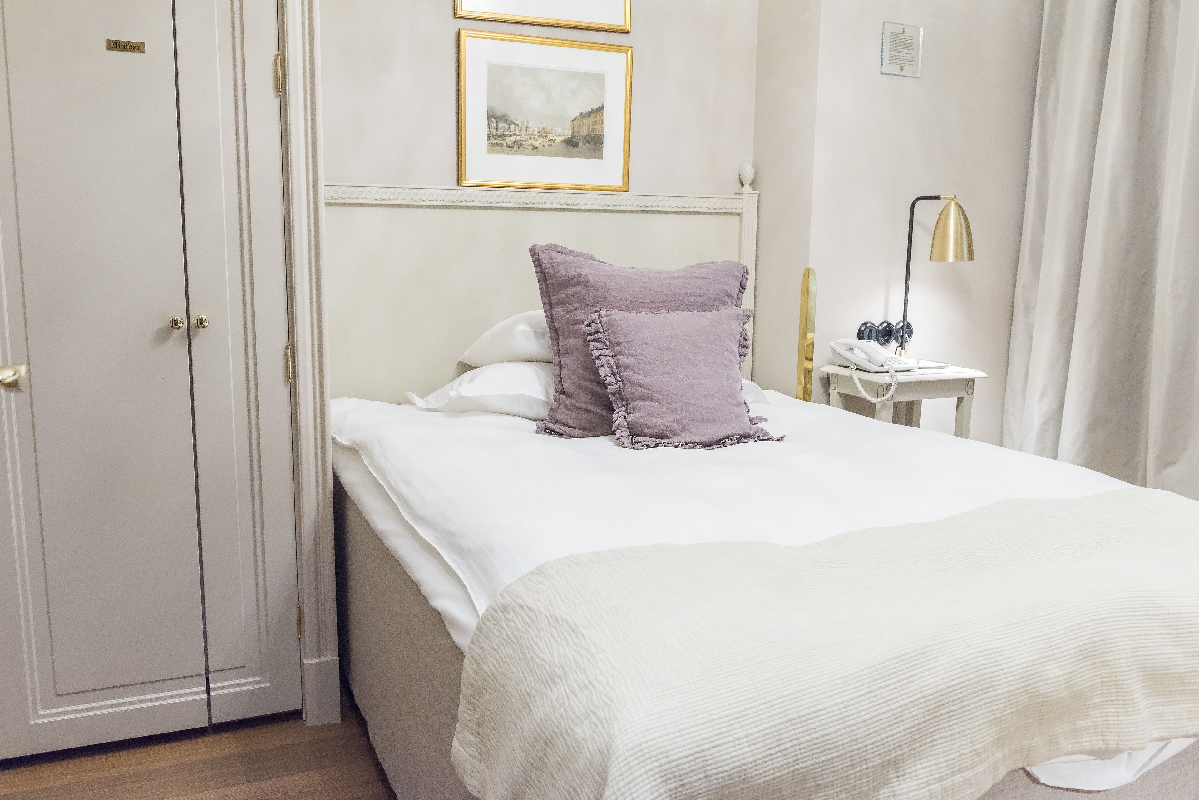 Een netjes opgemaakt bed met een wit en paars kussen, een nachtkastje met een lamp en een telefoon.