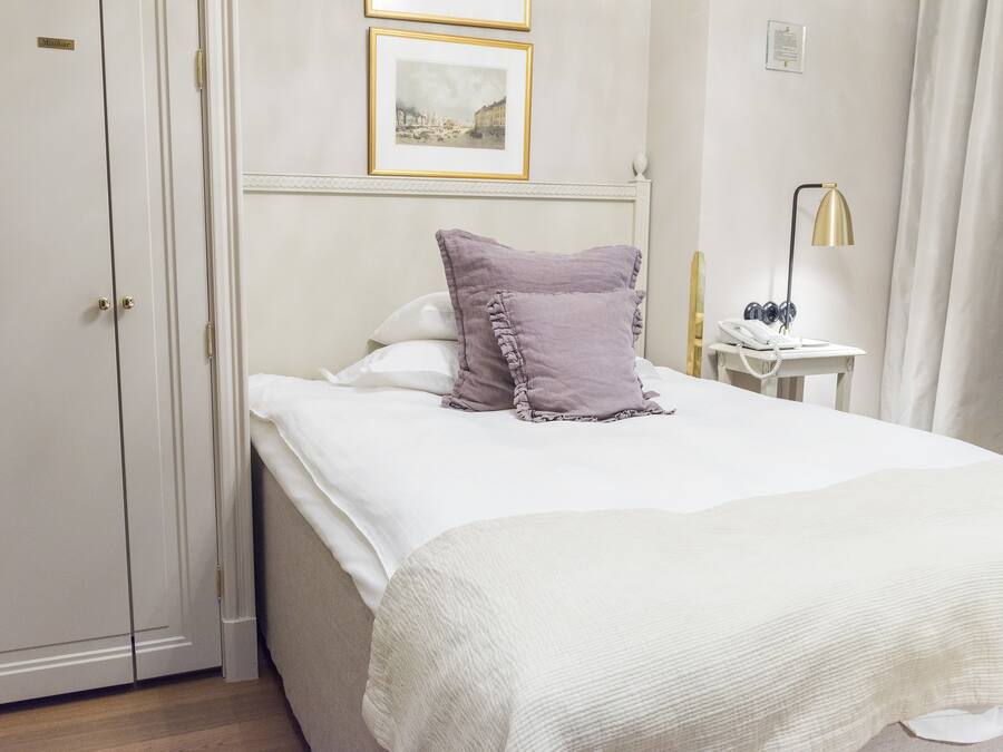 Een netjes opgemaakt bed met een wit en paars kussen, een nachtkastje met een lamp en een telefoon.