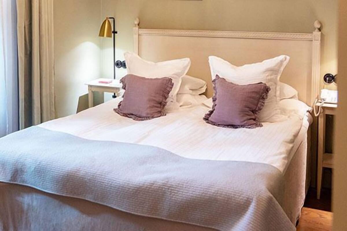 Een netjes opgemaakt bed in wit en roze, een nachtkastje met een lamp, en een raam met gordijnen.