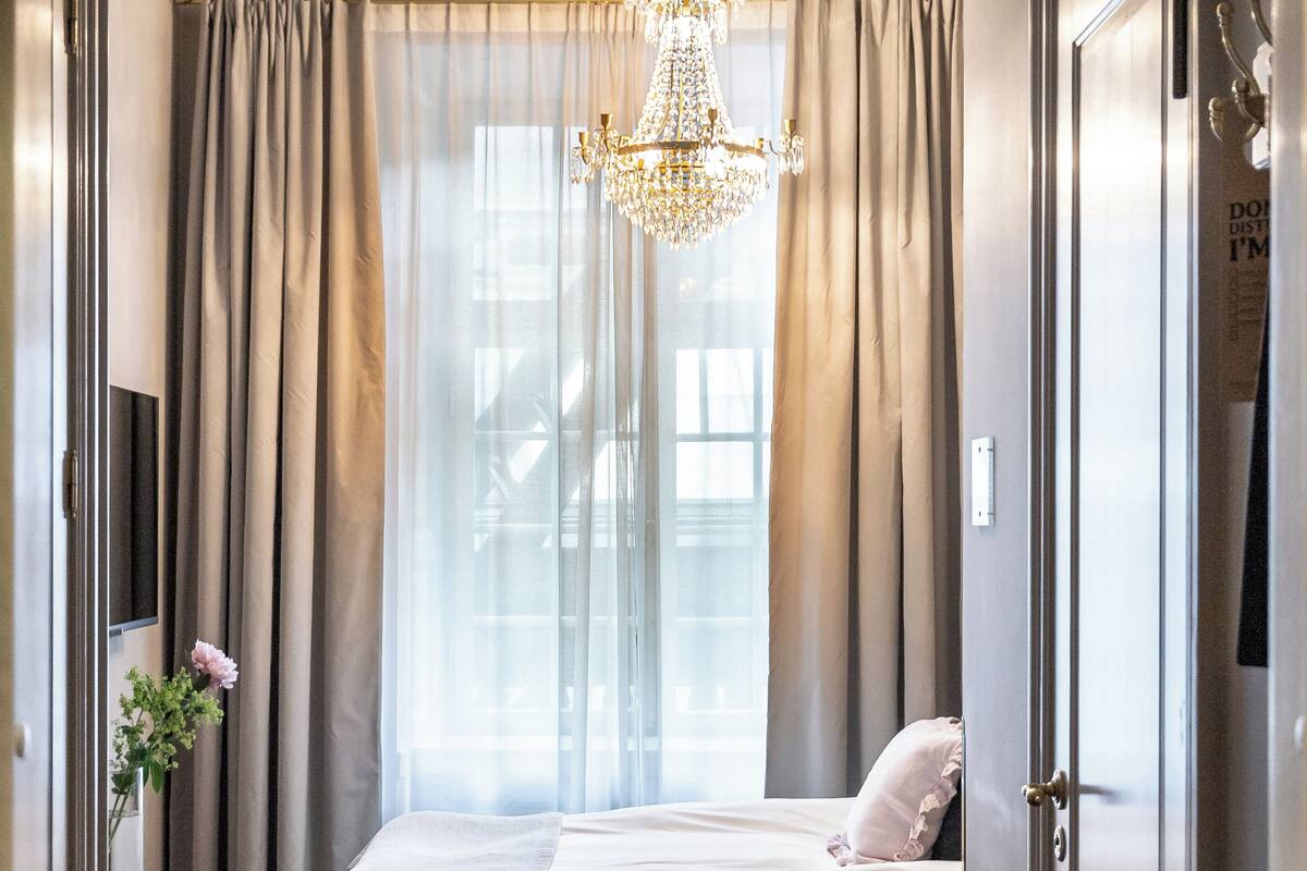 Een hotelkamer met een bed, gordijnen, een kroonluchter en uitzicht door het raam.