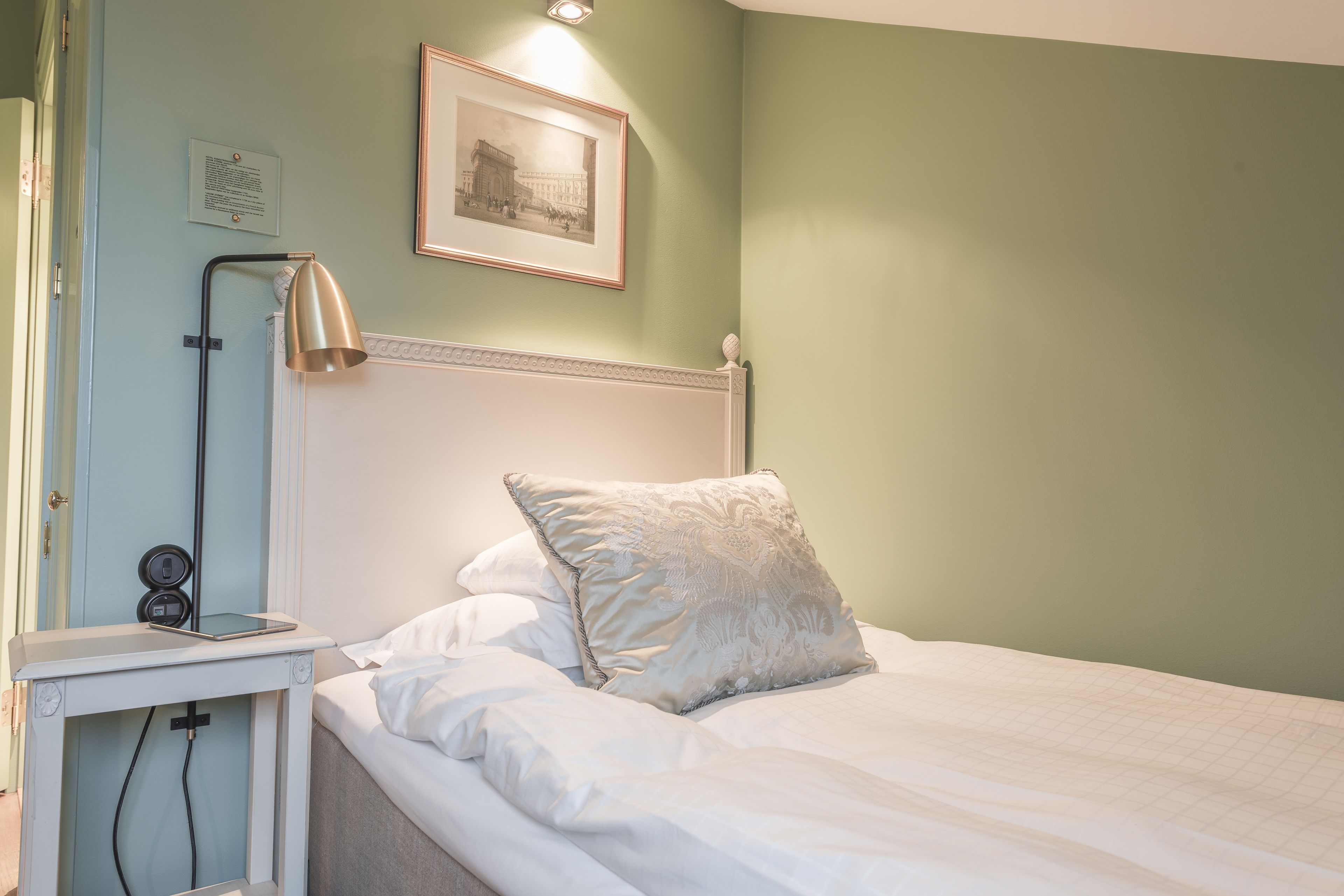 Una cama bien hecha con edredón blanco y almohada decorativa, una mesita de noche con lámpara y un cuadro enmarcado en la pared.