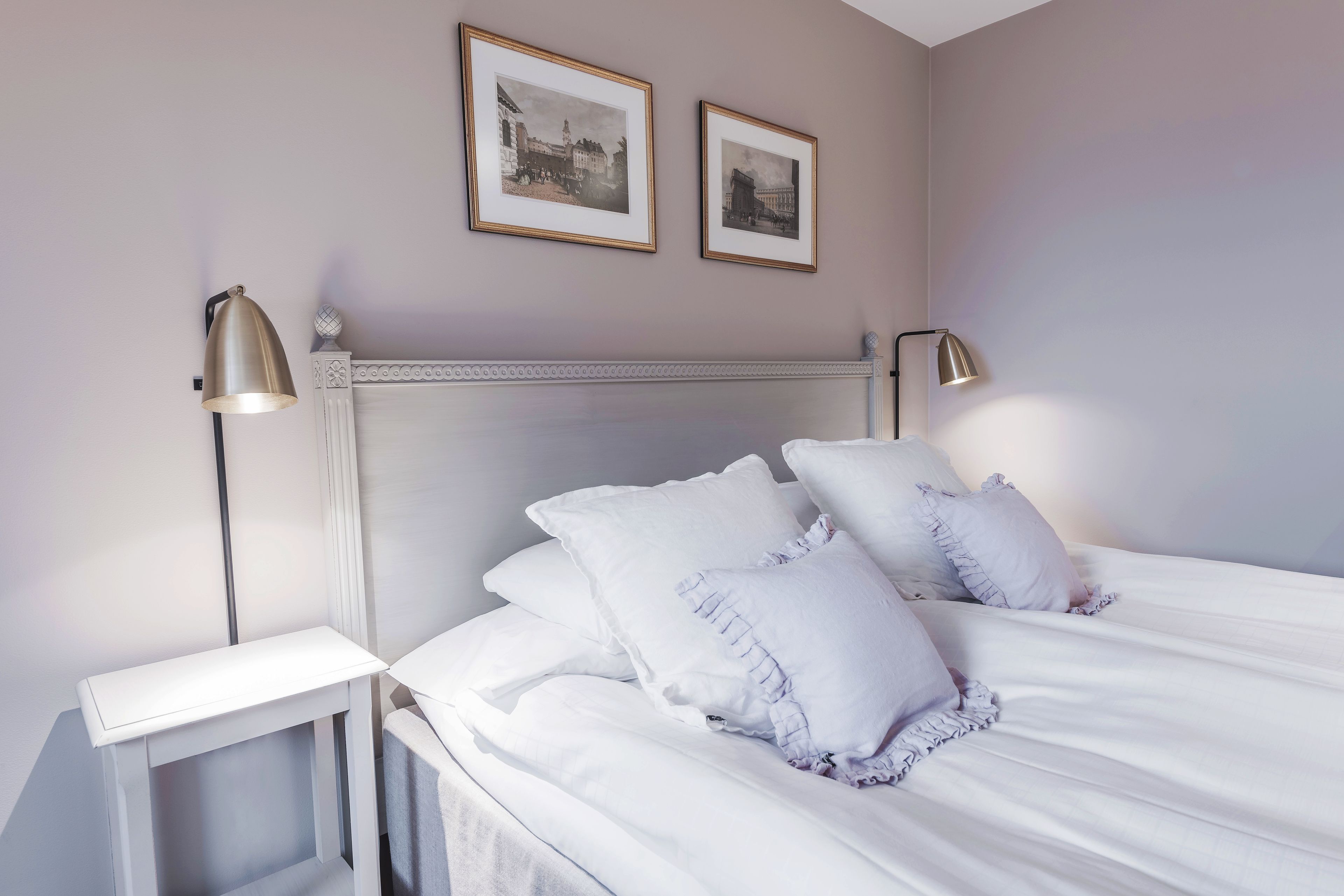A neatly made bed with white linens and ruffled pillows, a white bedside table, and two wall-mounted lamps.