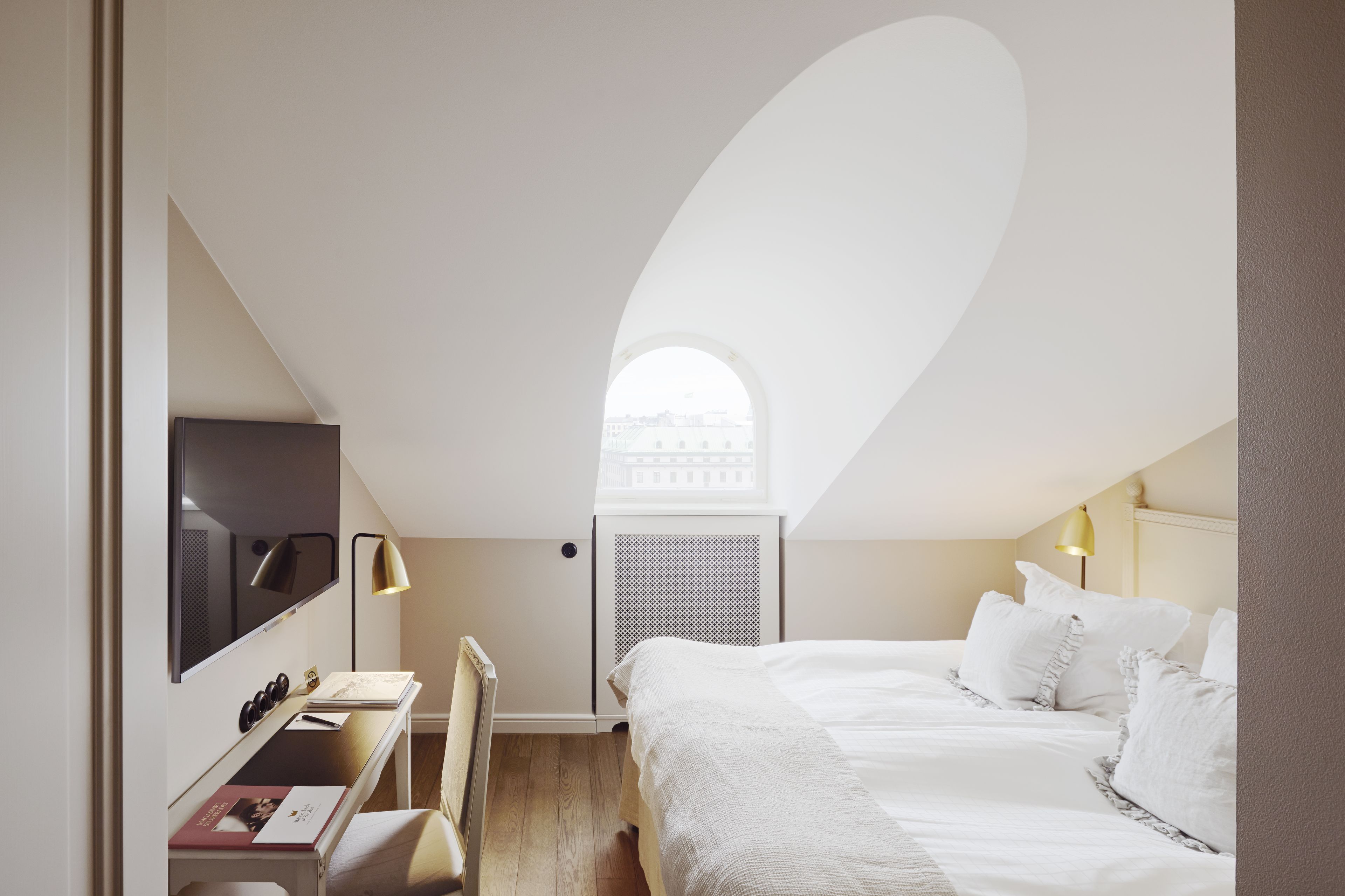 Een slaapkamer met een bed, een bureau, een stoel, een televisie en een raam met uitzicht op gebouwen.