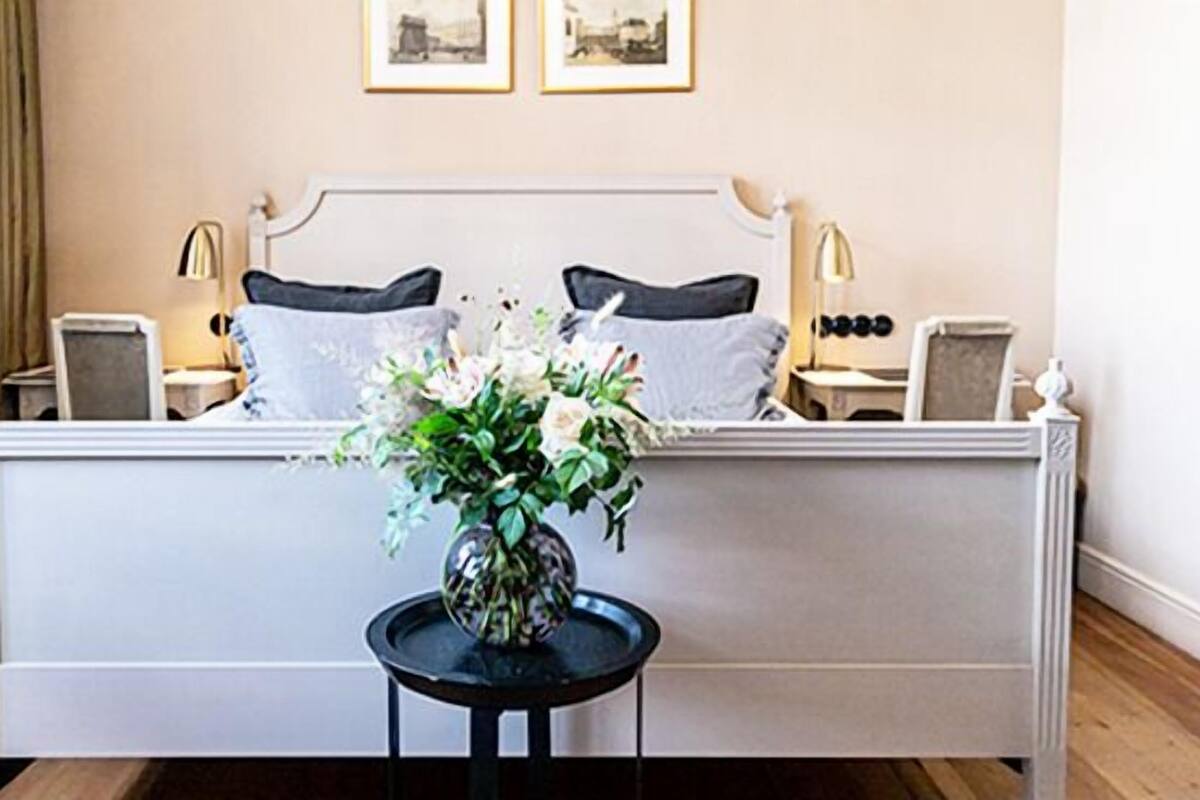 Een net opgemaakt bed met witte lakens, een vaas met bloemen op een bijzettafeltje en twee ingelijste foto's aan de muur.