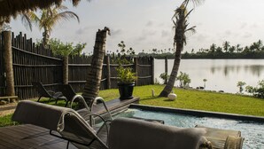 A pool area with lounge chairs, a wooden deck, and a view of a calm lake surrounded by palm trees.