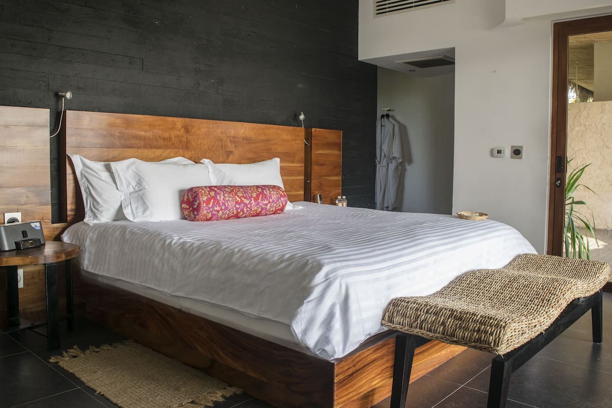 Un dormitorio moderno con cabecera de madera, una cama grande con ropa de cama blanca y a rayas, una mesita de noche con un radio y un banco tejido.