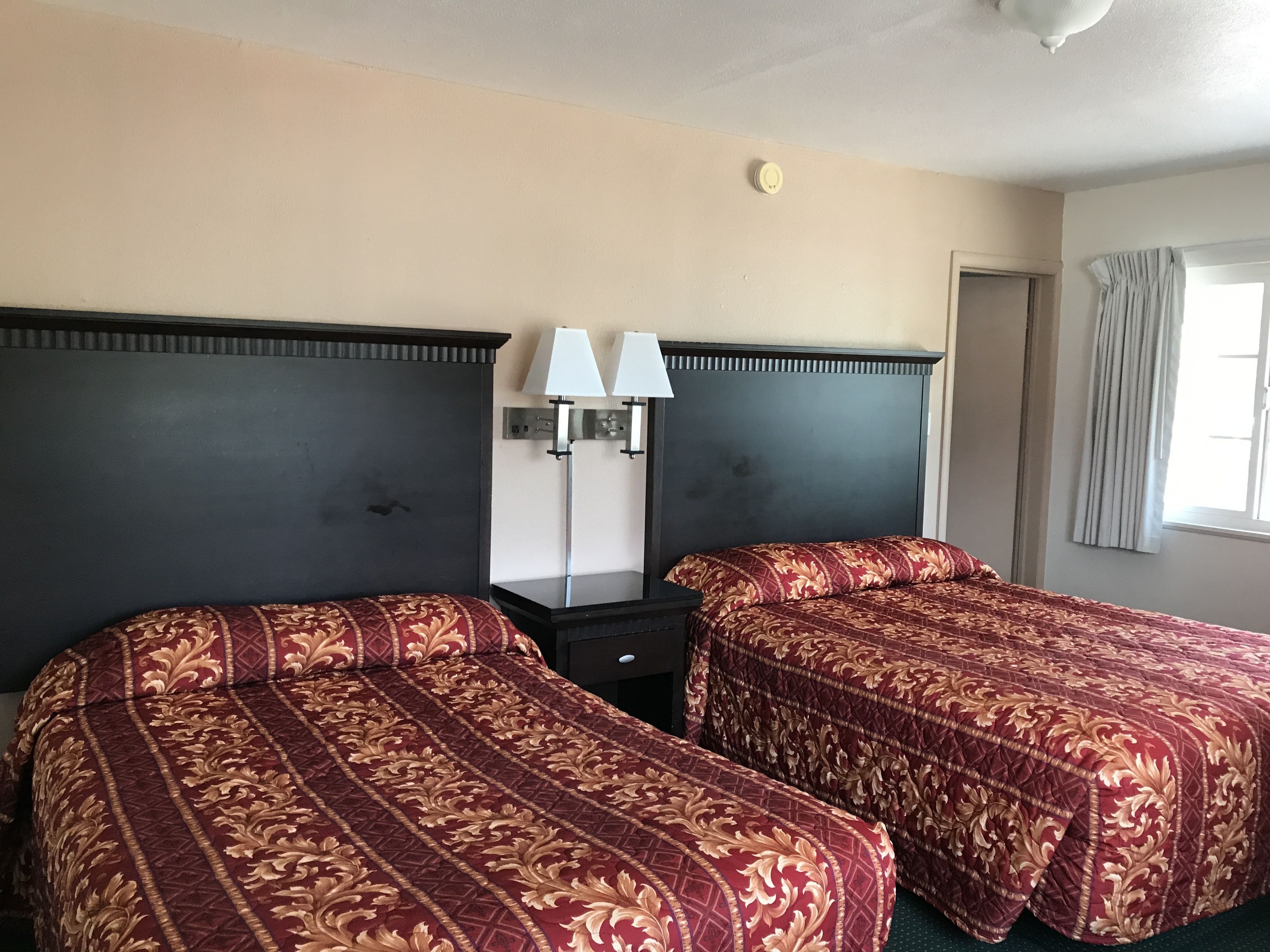 Habitación de hotel con dos camas, cada una con un cabecero oscuro y una colcha estampada roja. Entre las camas hay una mesita de noche con una lámpara, y a la derecha se ve una ventana con cortinas.