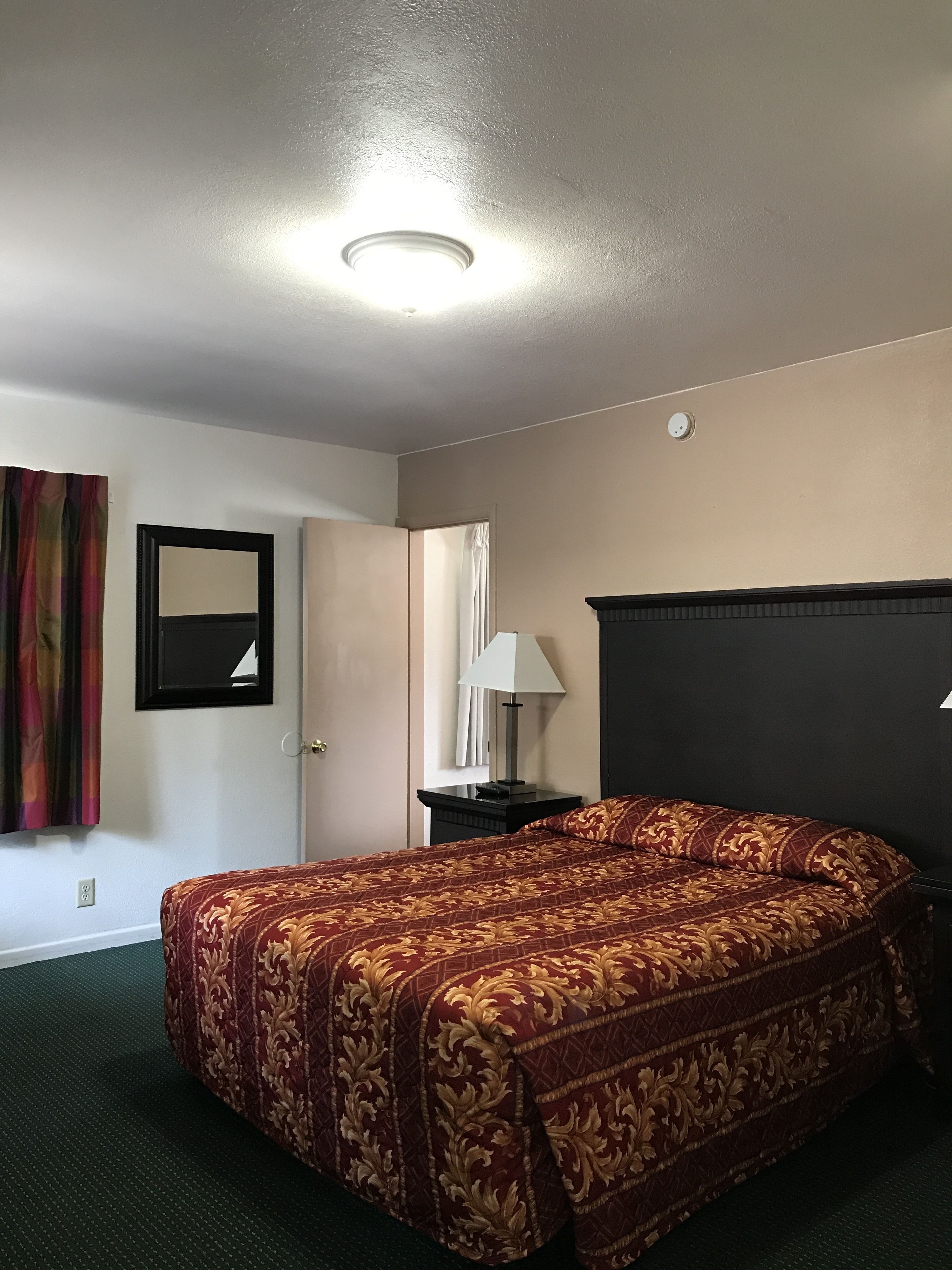 A hotel room with a large bed, a nightstand, a lamp, a mirror, and a door.