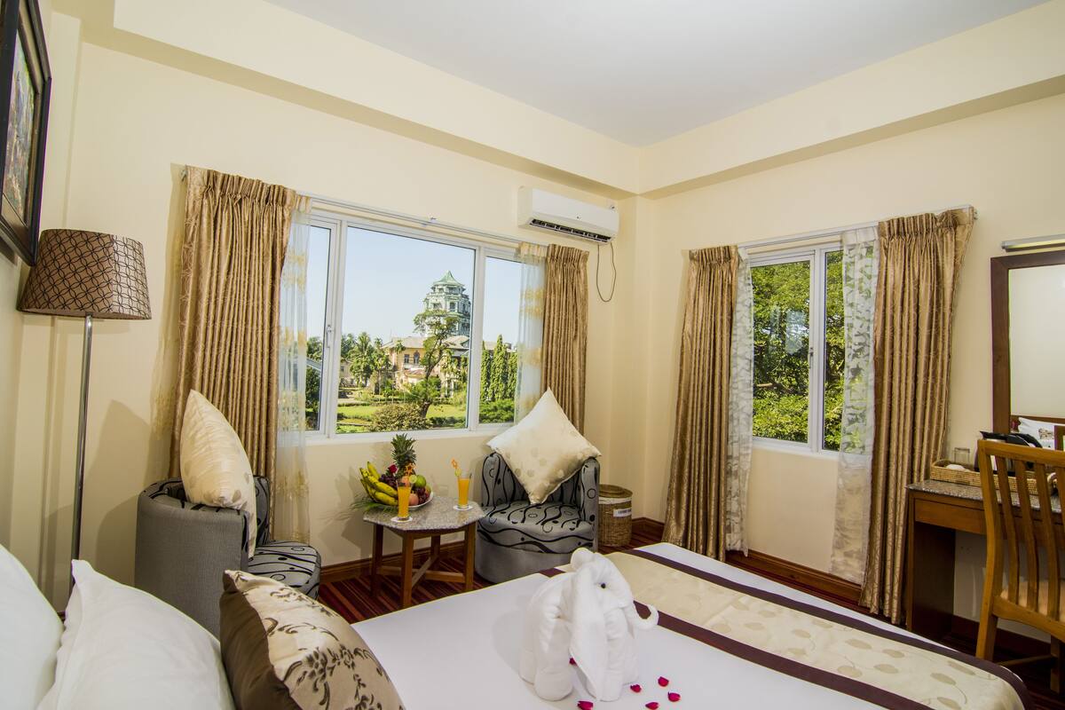 Una habitación de hotel con cama, escritorio, silla, mesa con frutas y una bebida, y cortinas.