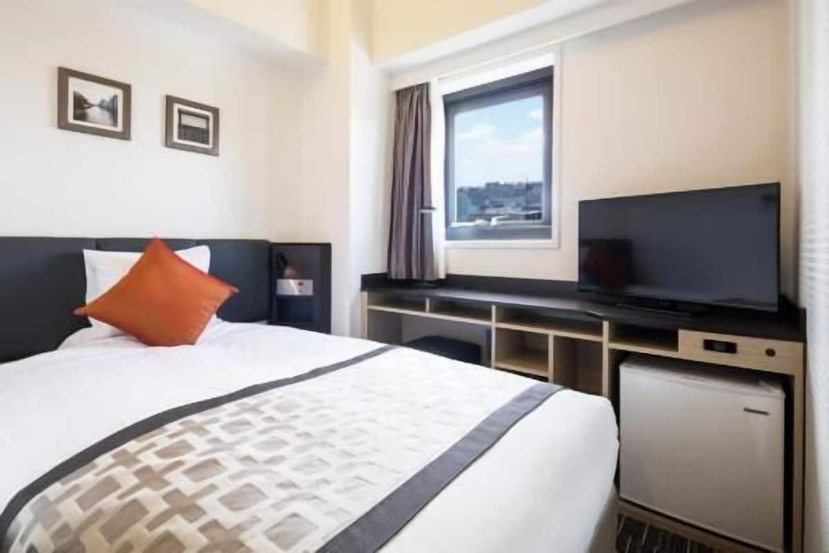A hotel room with a bed, a desk with a television, a window with curtains, and a small refrigerator.
