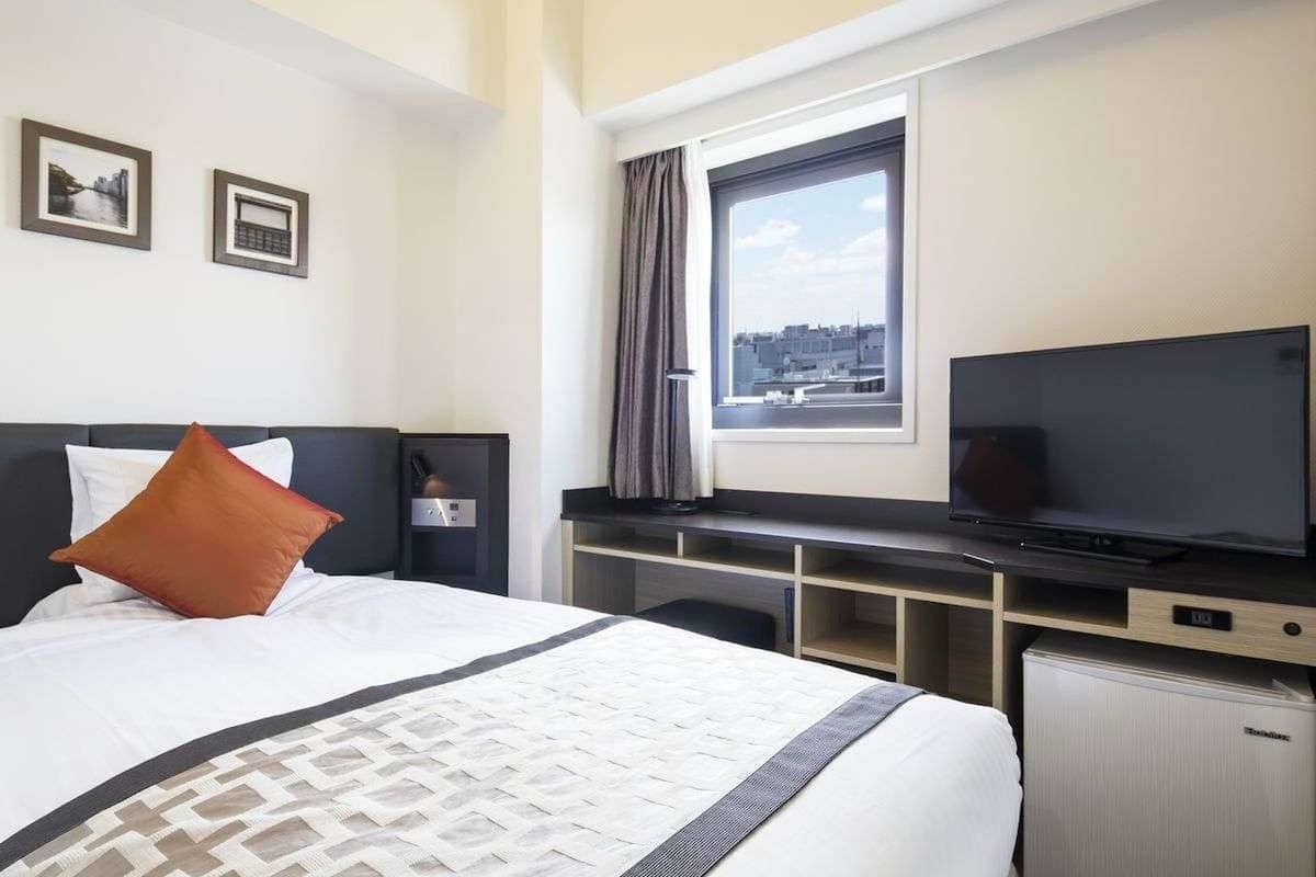 A hotel room with a bed, a desk with a flat-screen TV, a small refrigerator, and a window with curtains.
