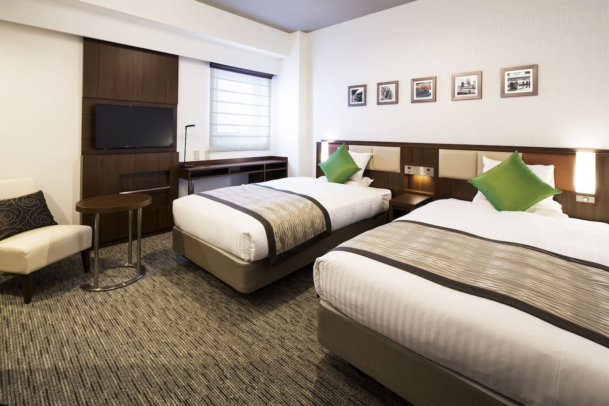 A hotel room with two beds, a TV mounted on the wall, a small table, and a bench.