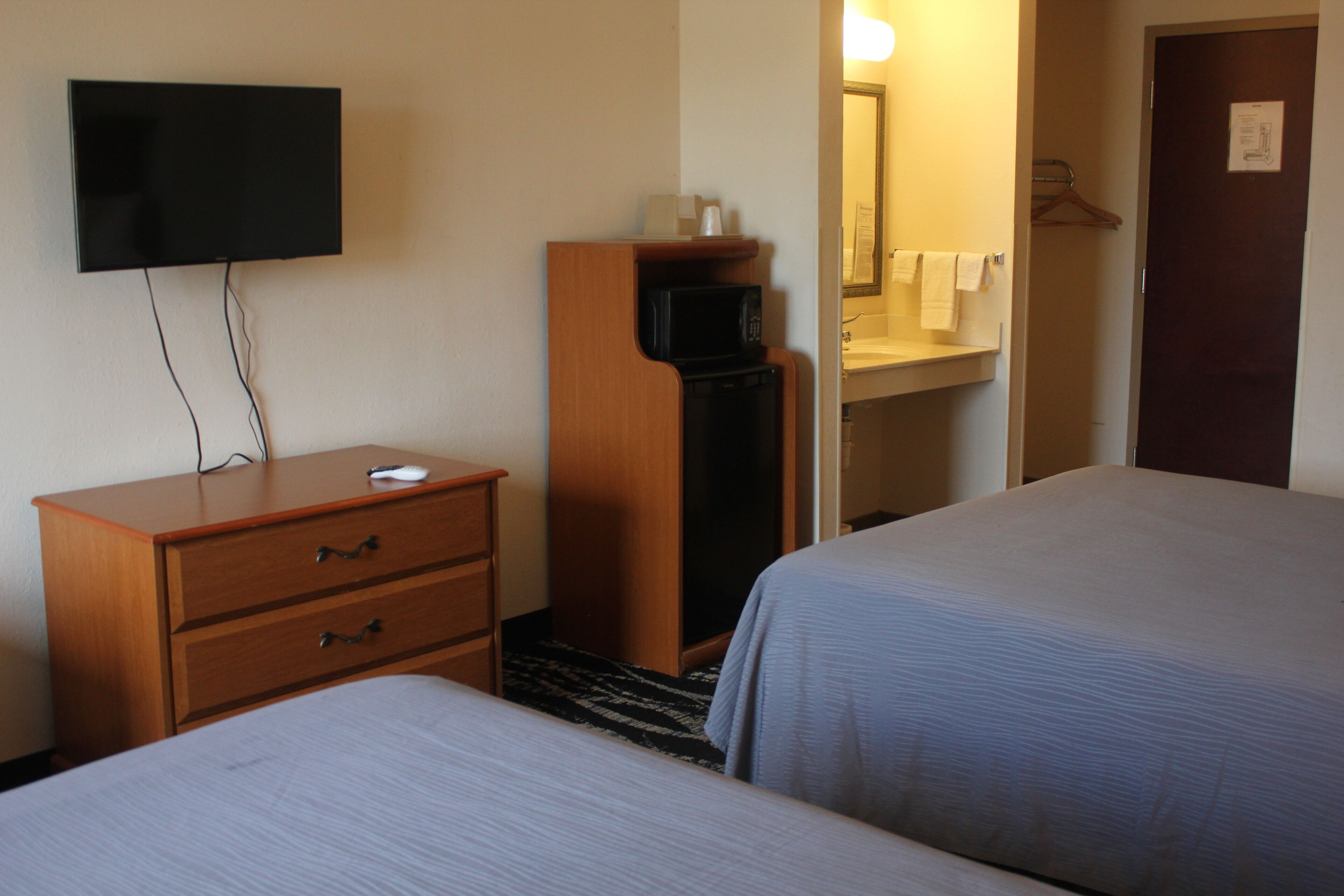 Une chambre d’hôtel avec deux lits, un téléviseur fixé au mur, une commode en bois, un four à micro-ondes et une salle de bain avec un miroir et un porte-serviettes.