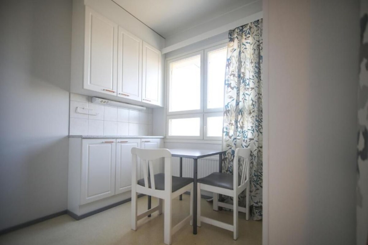 Een kleine keuken met witte kasten, een raam met gordijnen en een tafel met stoelen.
