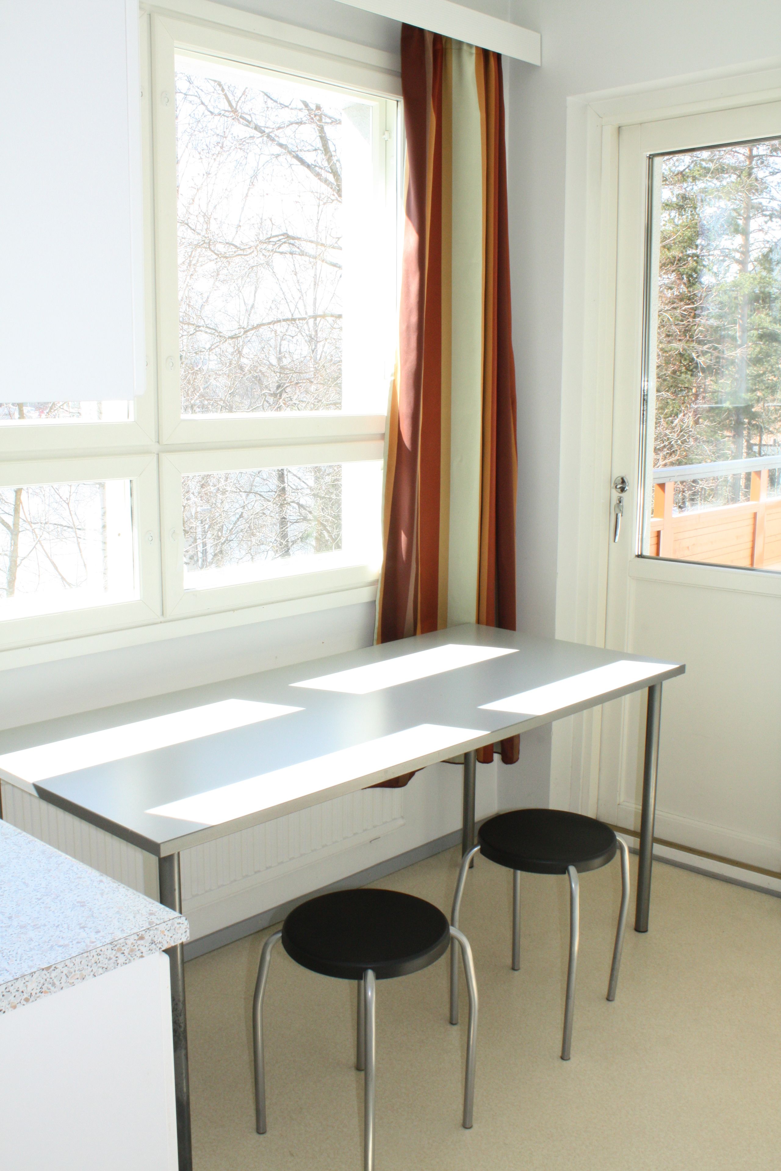 A room with a white desk, two black chairs, a window with curtains, and a door.