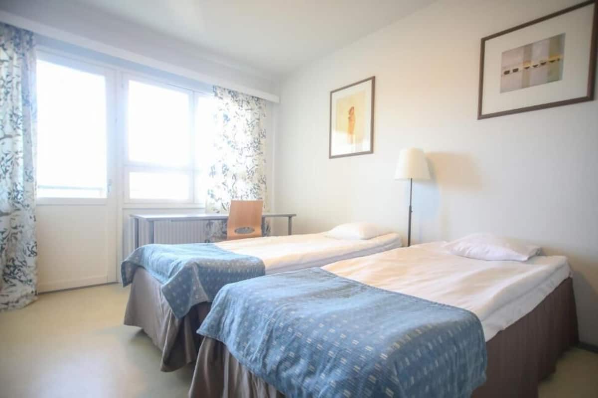 A hotel room with two beds, a desk, a chair, a lamp, and two framed pictures on the wall.