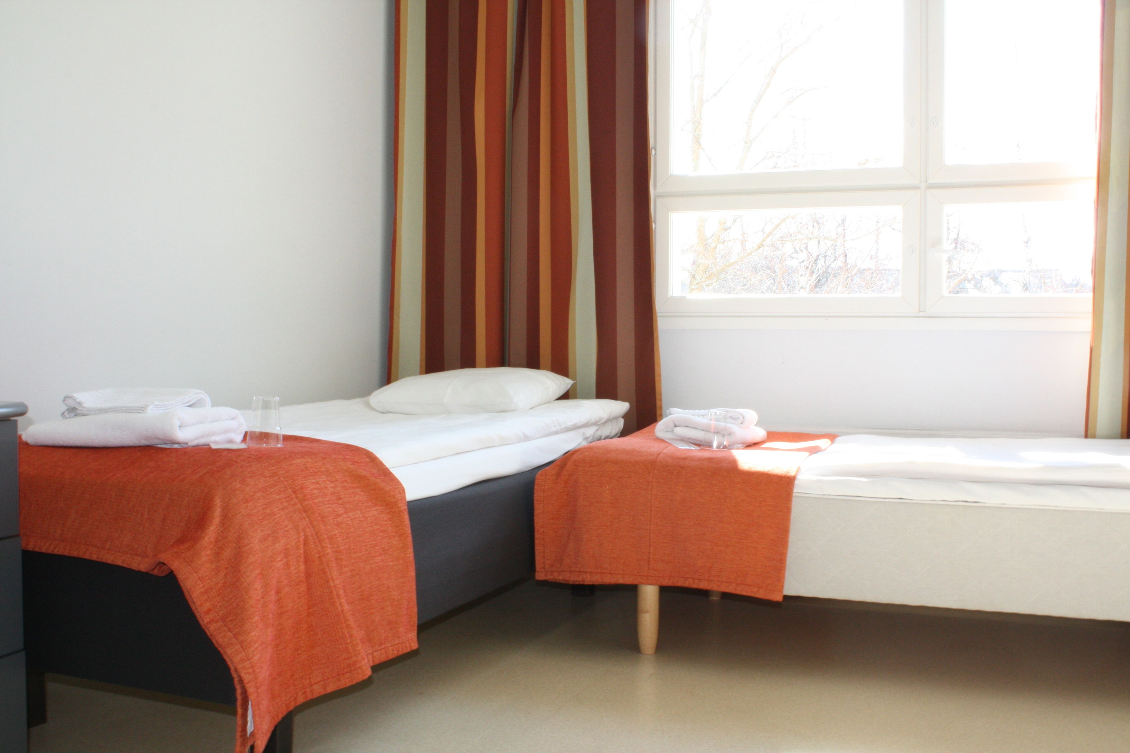 A hotel room with two single beds, each with a white mattress and orange bedspread. There is a window with curtains, and a small table with a glass of water and a towel on it.