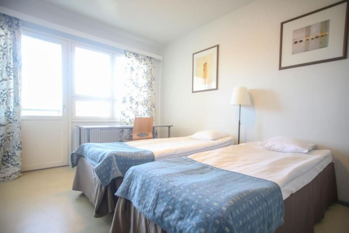 Habitación de hotel con dos camas, un escritorio, una silla, una lámpara y dos cuadros en la pared.