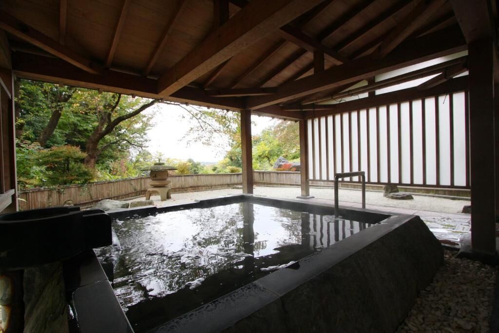 A hot spring area with a wooden pergola, stone seating, and a view of the surrounding greenery.