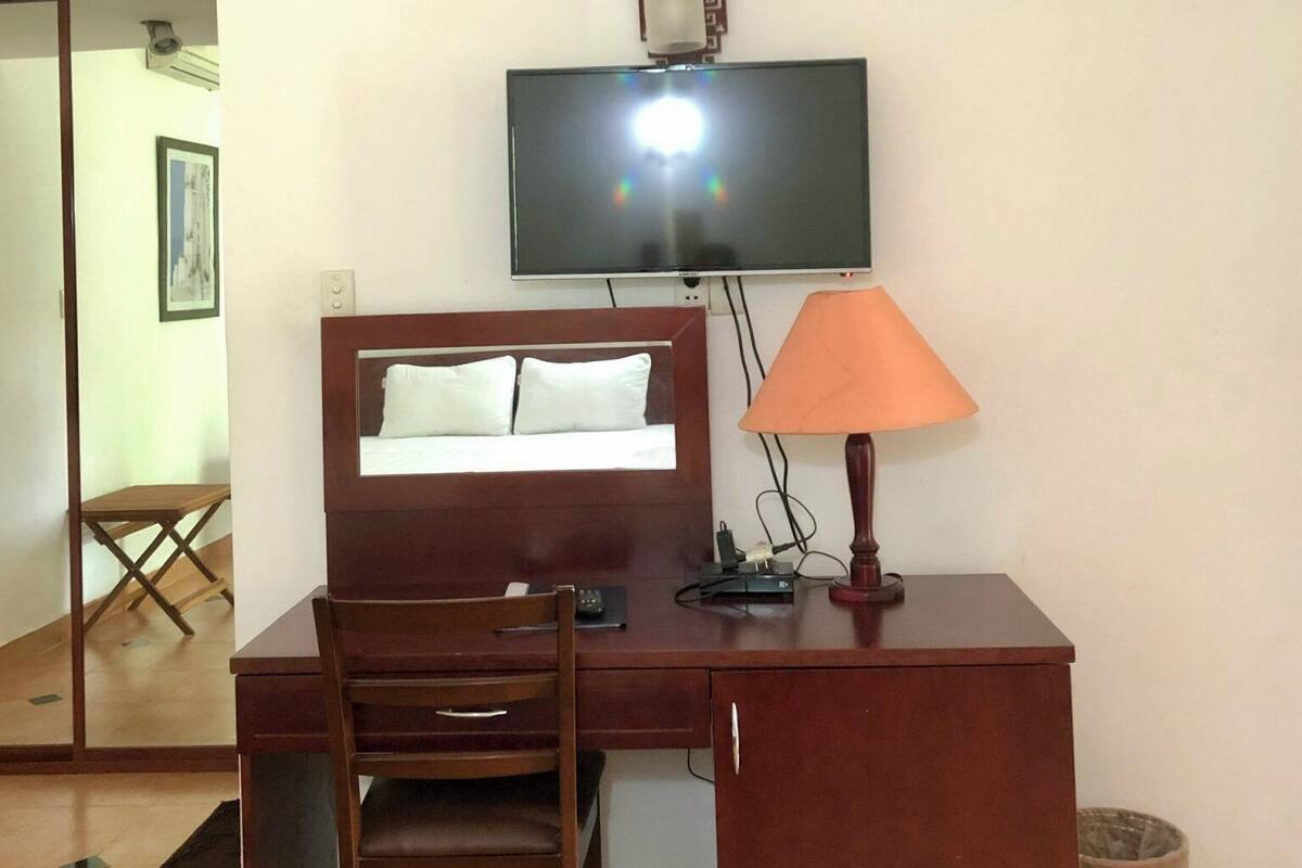 Hotellihuone, jossa on puinen työpöytä, tuoli, sänky, seinälle asennettu televisio ja lamppu.