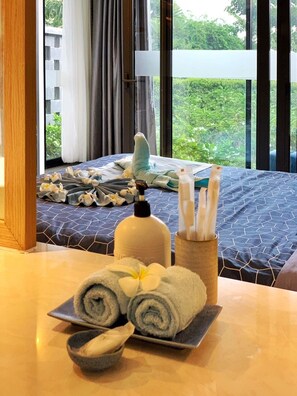 Habitación de hotel con cama, mesa con toallas, artículos de tocador y vistas a la vegetación.