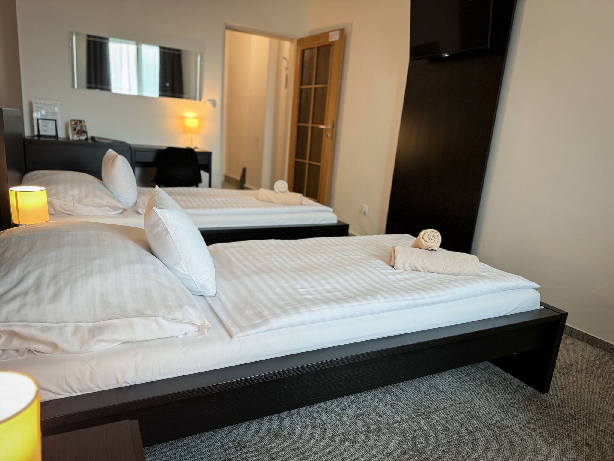 Quarto de hotel com duas camas, uma escrivaninha, uma cadeira, uma luminária e uma televisão.