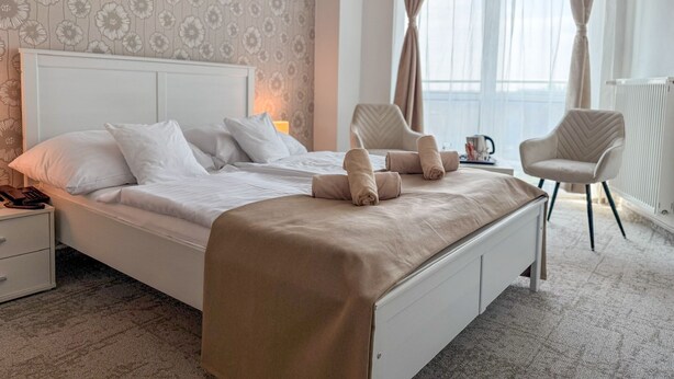 A neatly made bed with pillows and a blanket, a bedside table with a lamp, two chairs, and a window with curtains. - Hotel Modena (Bratislava)