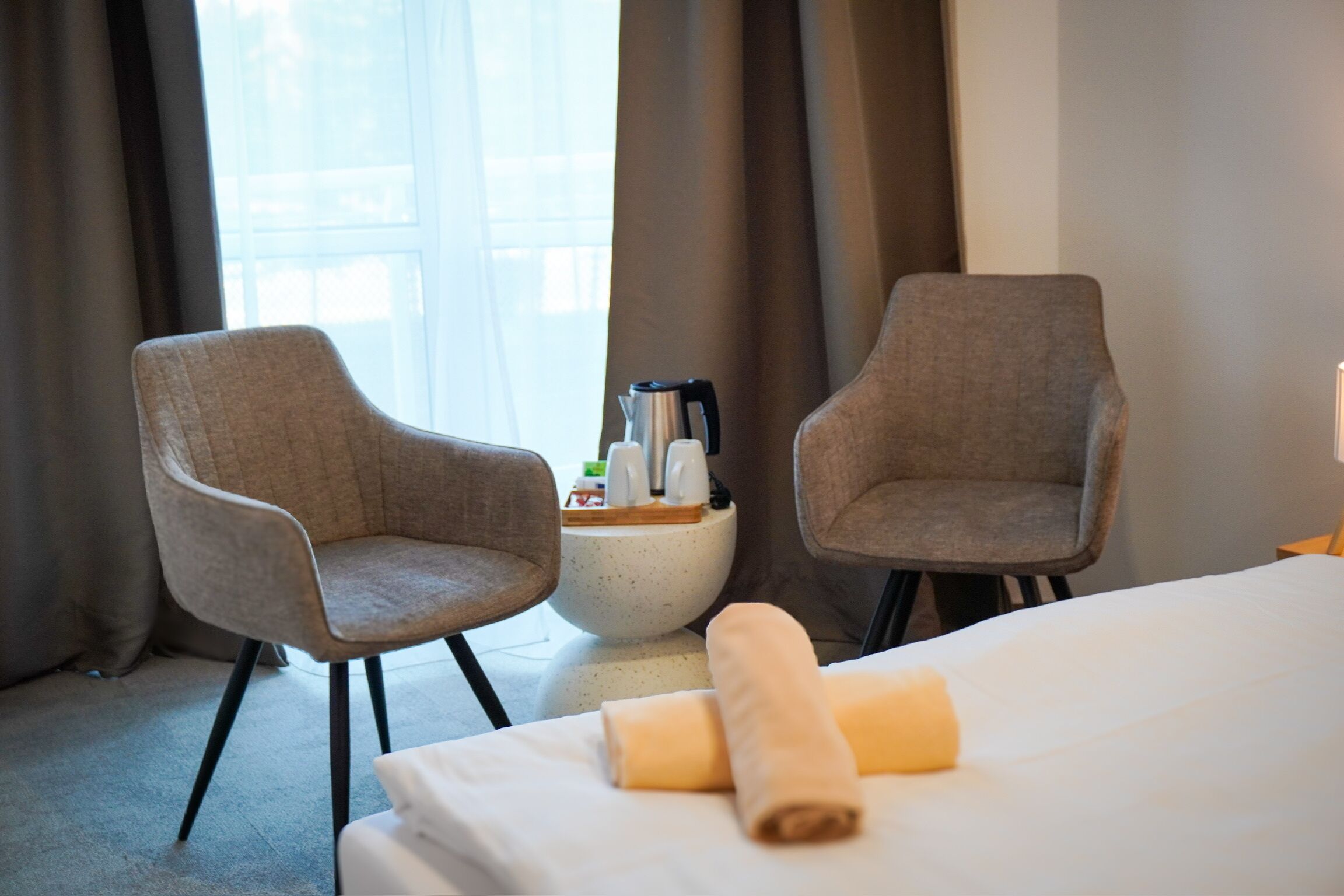 Een hotelkamer met twee grijze fauteuils, een bed met een witte lakenset en opgerolde handdoeken, een kleine ronde tafel met een waterkoker en kopjes, en een raam met gordijnen.