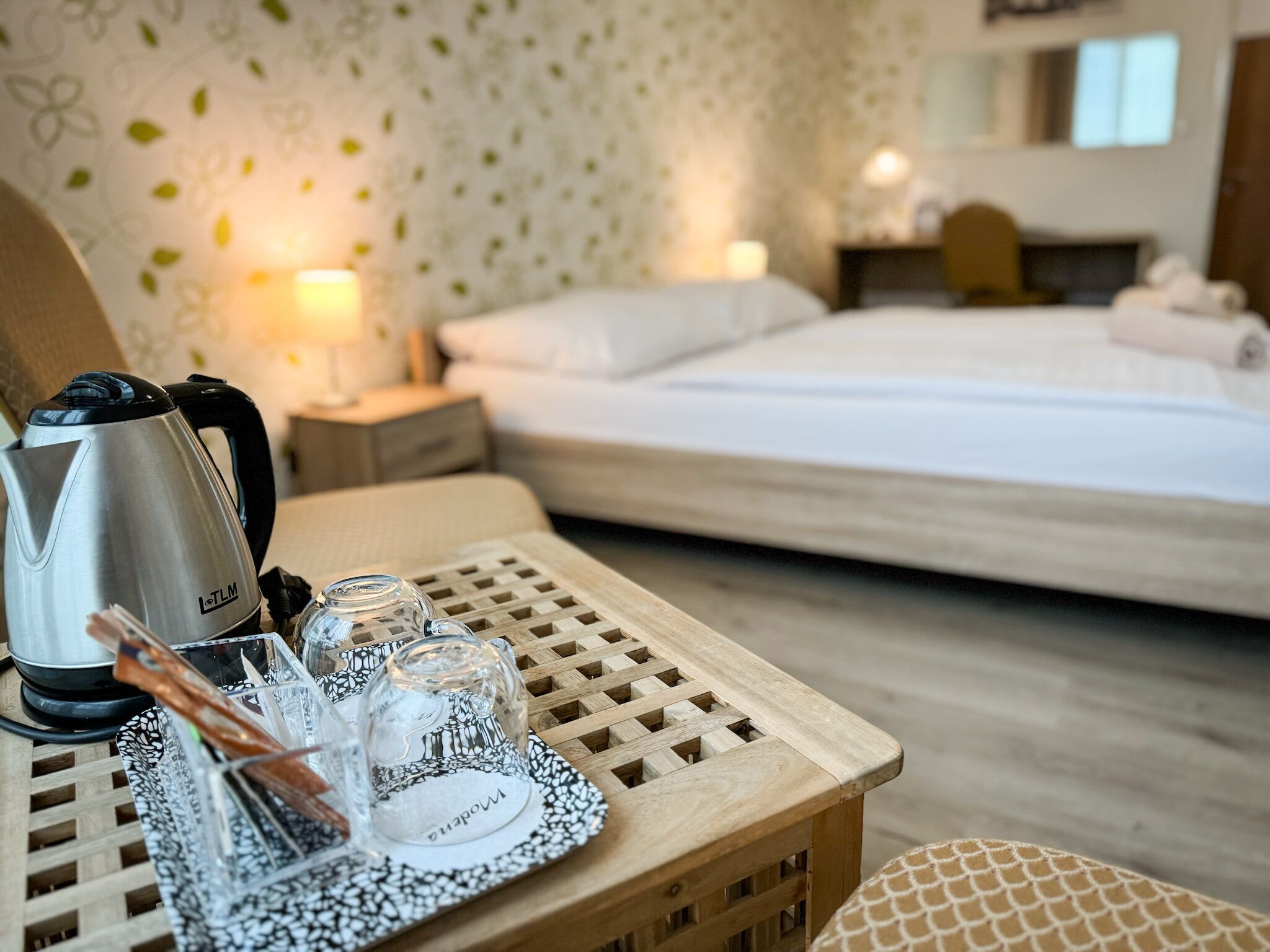 Ein Hotelzimmer mit einem Bett, einem Holztisch mit Wasserkocher, Gläsern und einem Teller.