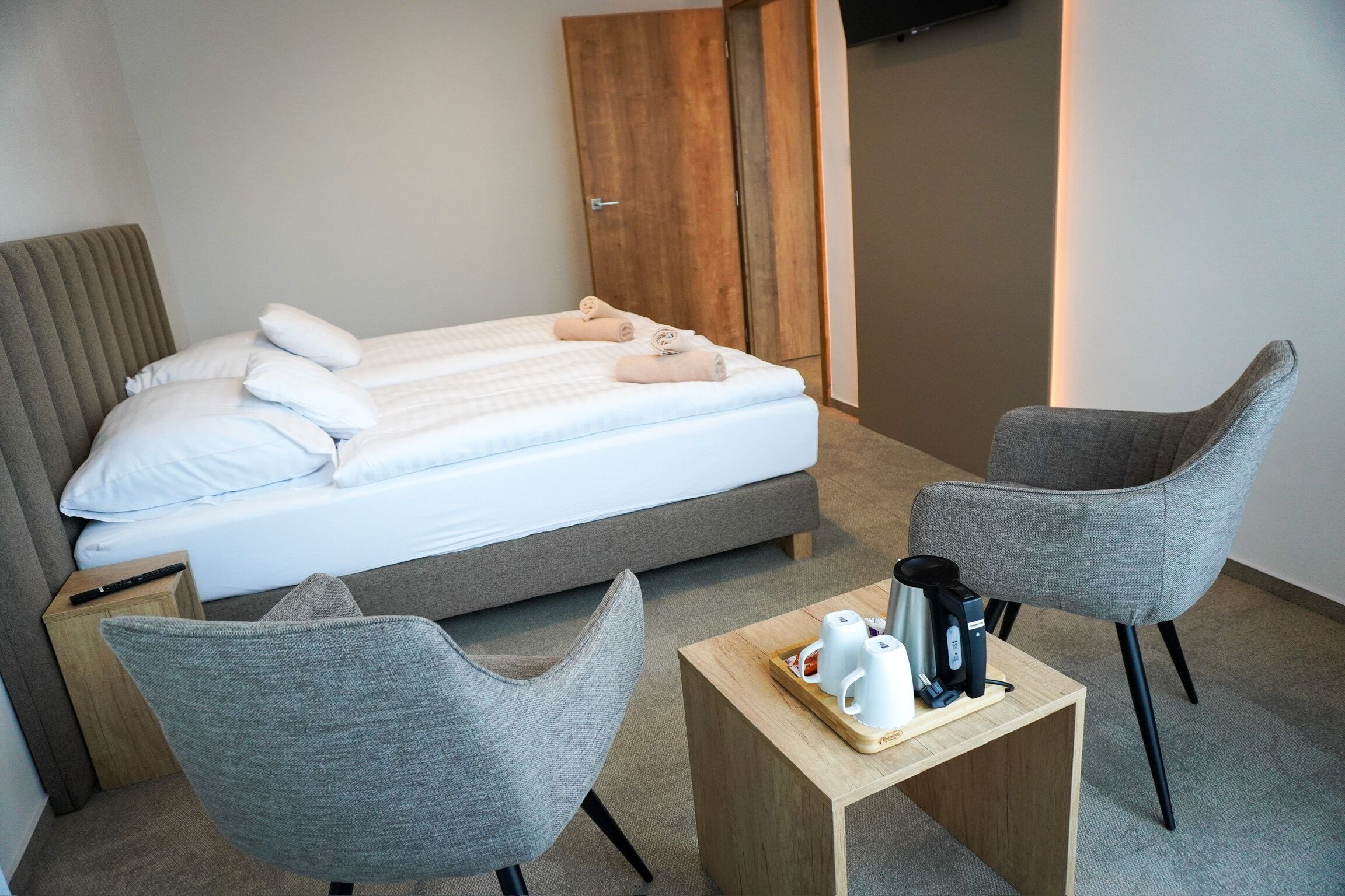 Hotellihuoneessa on sänky, kaksi harmaata nojatuolia, pieni puinen pöytä, jossa on kahvinkeitin, ja vaatekaappi.