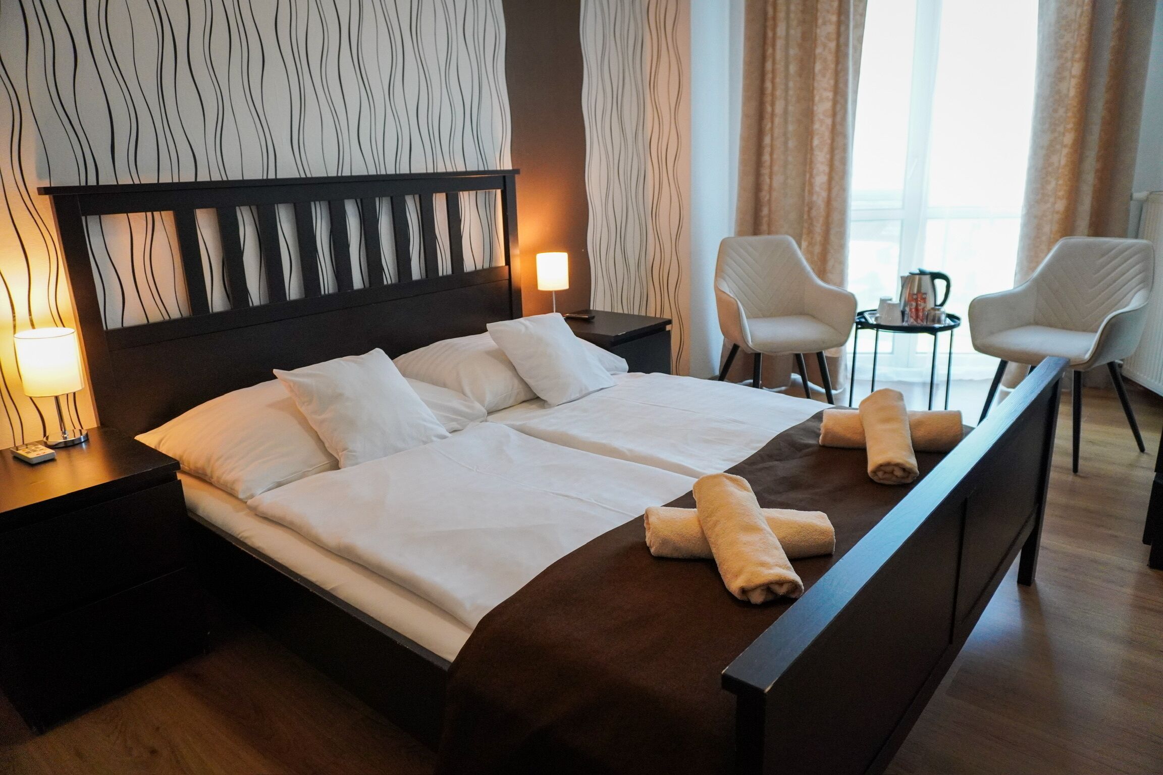 Habitación de hotel con una cama grande, dos mesitas de noche, un escritorio con sillas y vistas al exterior a través de cortinas.