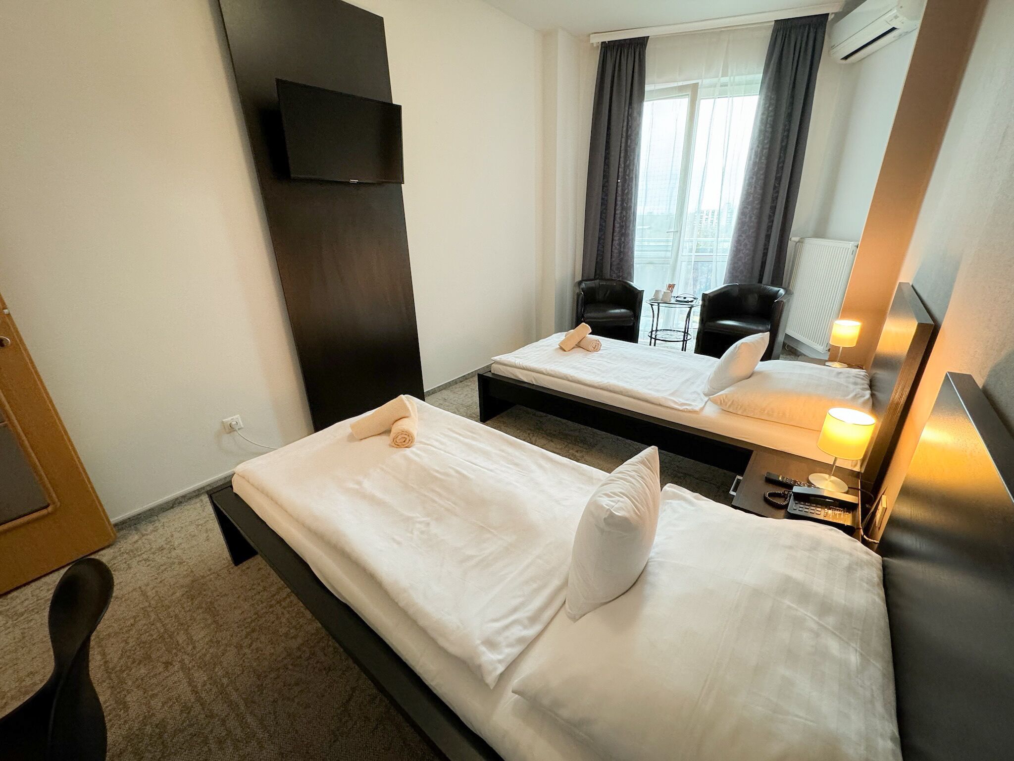 A hotel room with two single beds, a TV mounted on the wall, a small table with chairs, and a window with curtains.