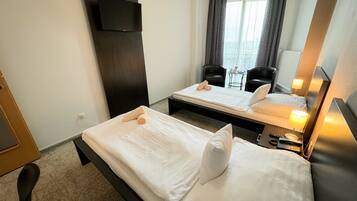 A hotel room with two single beds, a TV mounted on the wall, a small table with chairs, and a window with curtains.